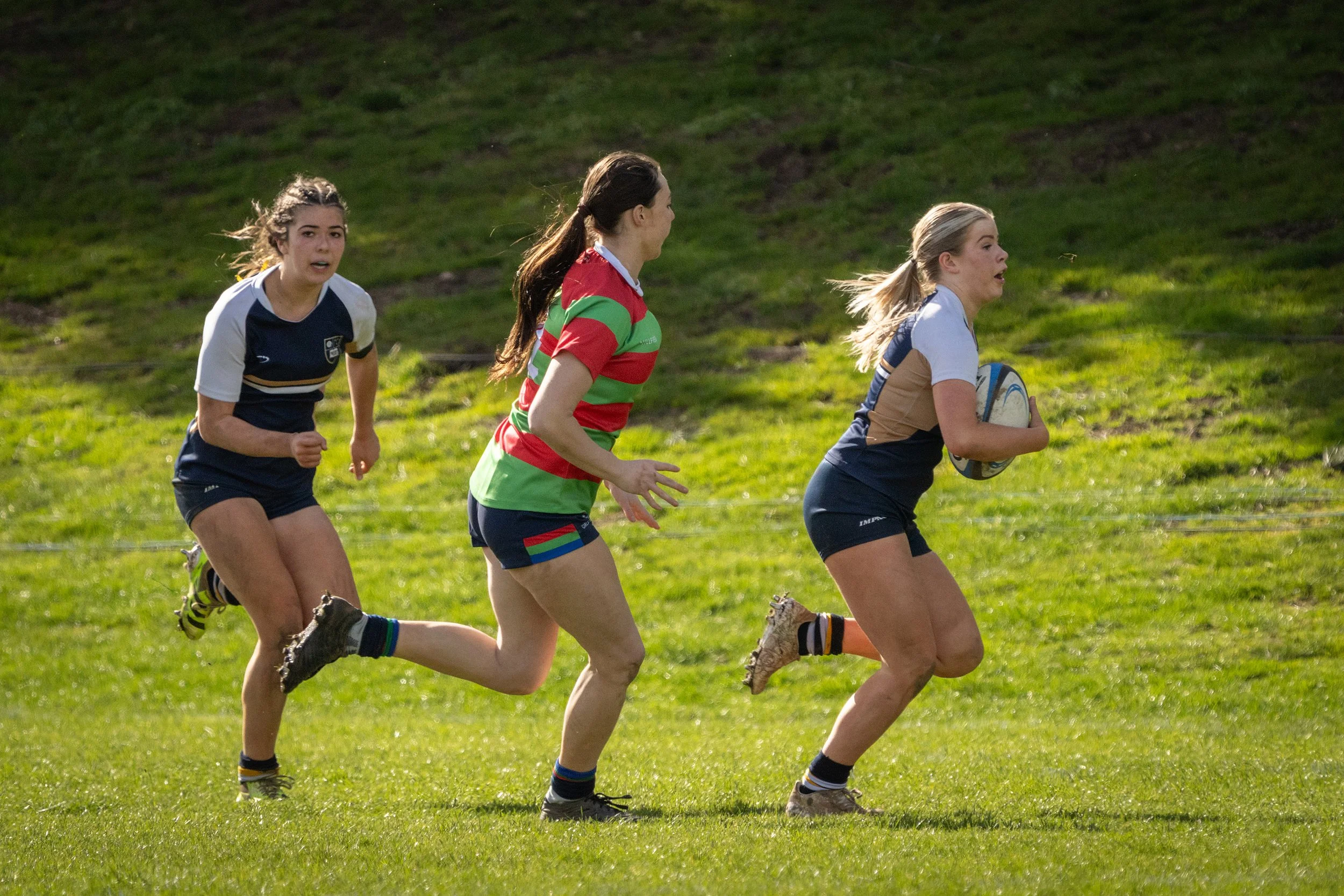 Three young women playing rugby on a grassy field. The woman in front, holding a rugby ball, wears a rugby uniform with dark shorts and a navy top. The woman in the middle is in a green and red striped jersey, while the woman on the left is in a navy