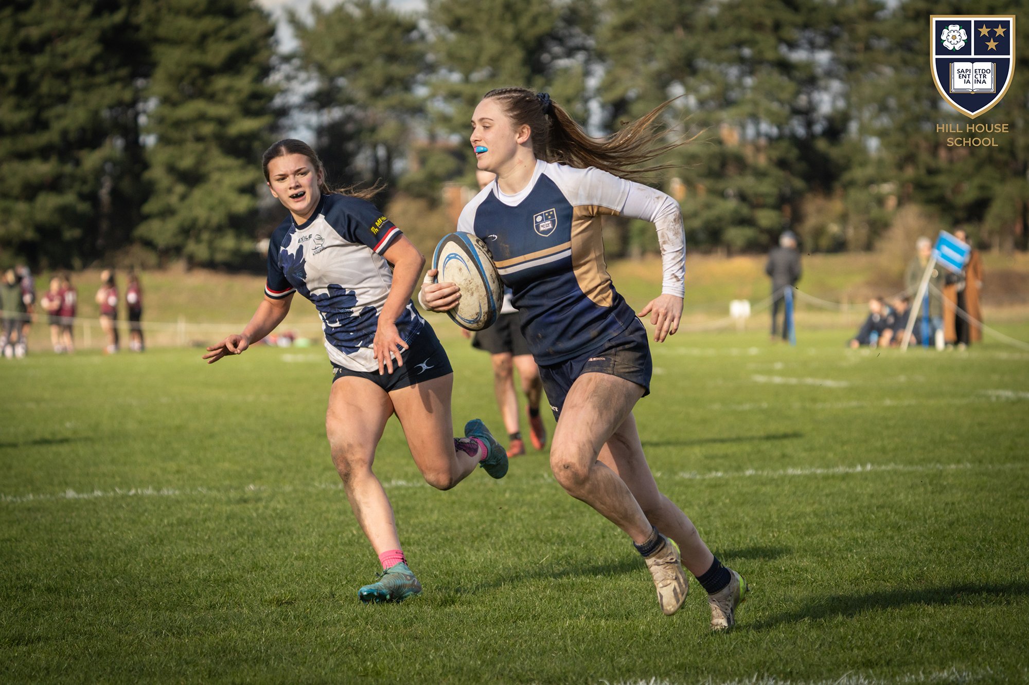 Two female rugby players competing for the ball on a grassy field during a match. One player is holding the rugby ball, and both are running. The players are wearing sports uniforms with logos. In the background, there are spectators and trees, and a