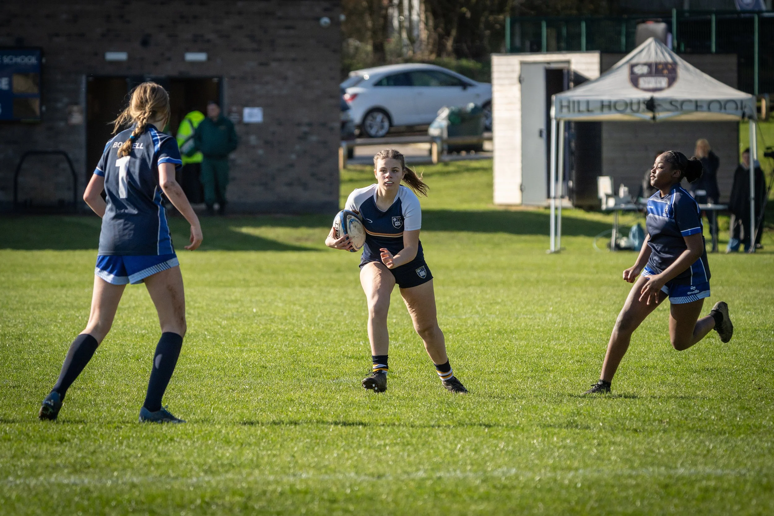 Young female rugby player with light skin, wearing a black and white uniform, holding a rugby ball, running on a grassy field during a game. Two other female players with dark hair in blue uniforms are nearby, and there are spectators, a tent, a car,