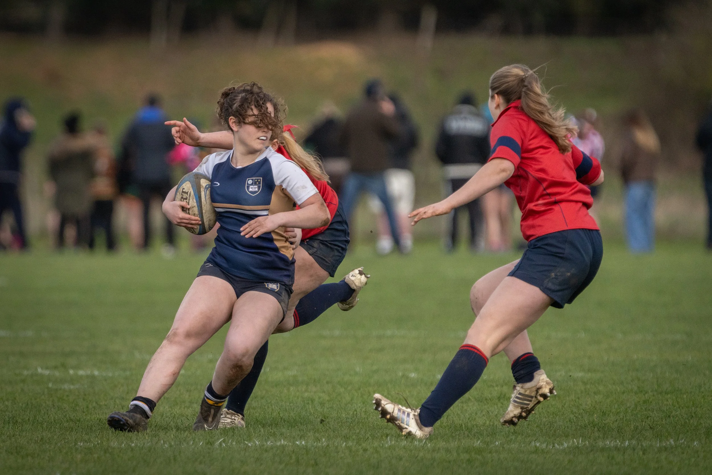 Girls playing rugby on a grassy field, with one girl holding a rugby ball and two others reaching toward her, blurred spectators in the background.