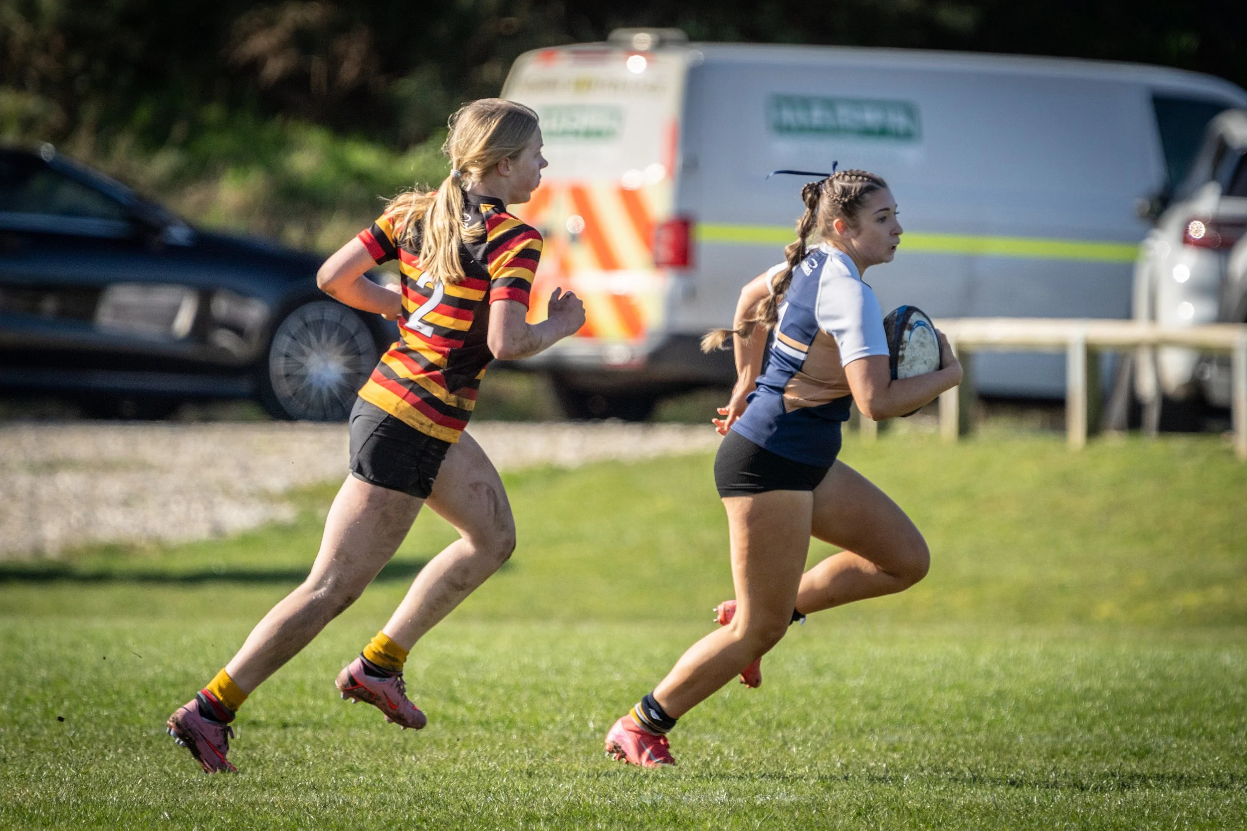 Two young women running on a grassy field, one holding a rugby ball, in athletic clothing.