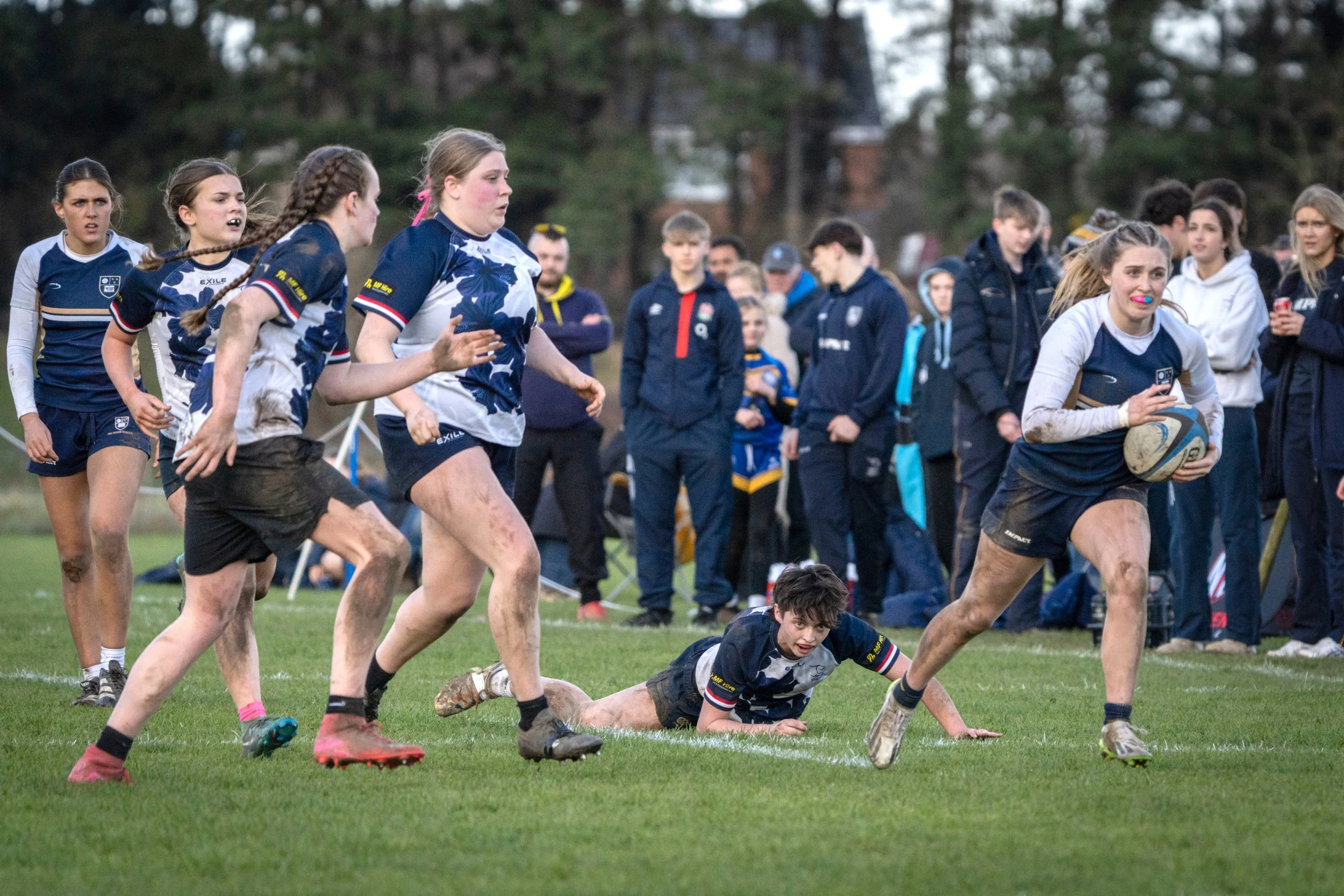 A group of female rugby players on a grassy field during a match. One player in white and blue is holding the ball while another in blue and white is running beside her. Several players and spectators are in the background, some watching and others e