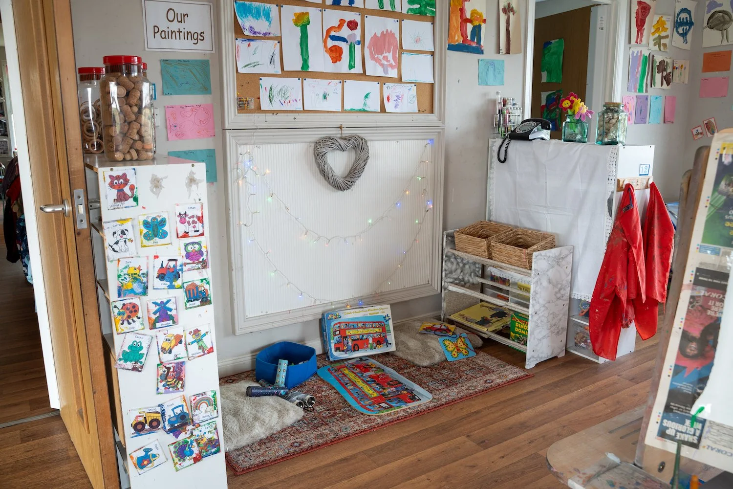 Children's artwork and crafts display area with colorful paintings, drawings, and craft projects, featuring a white sign labeled 'Our Paintings,' a ABC card display, some toys, wicker baskets, and decorative lights.