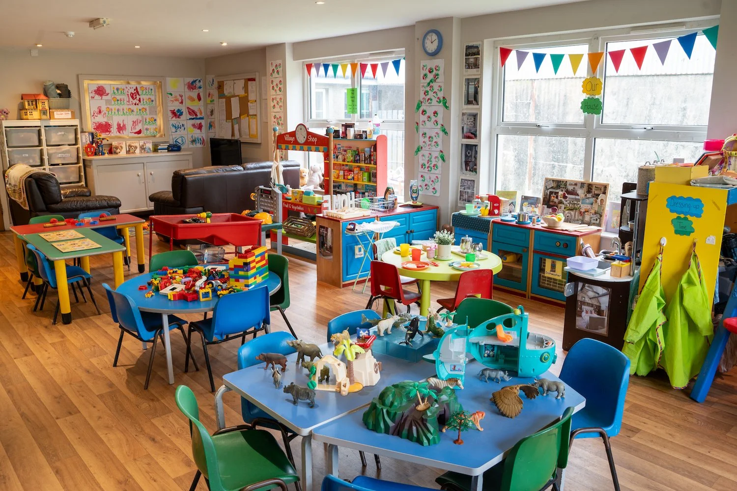 Colorful children's playroom filled with tables, chairs, toys, and educational materials, decorated with bunting and children's artwork.