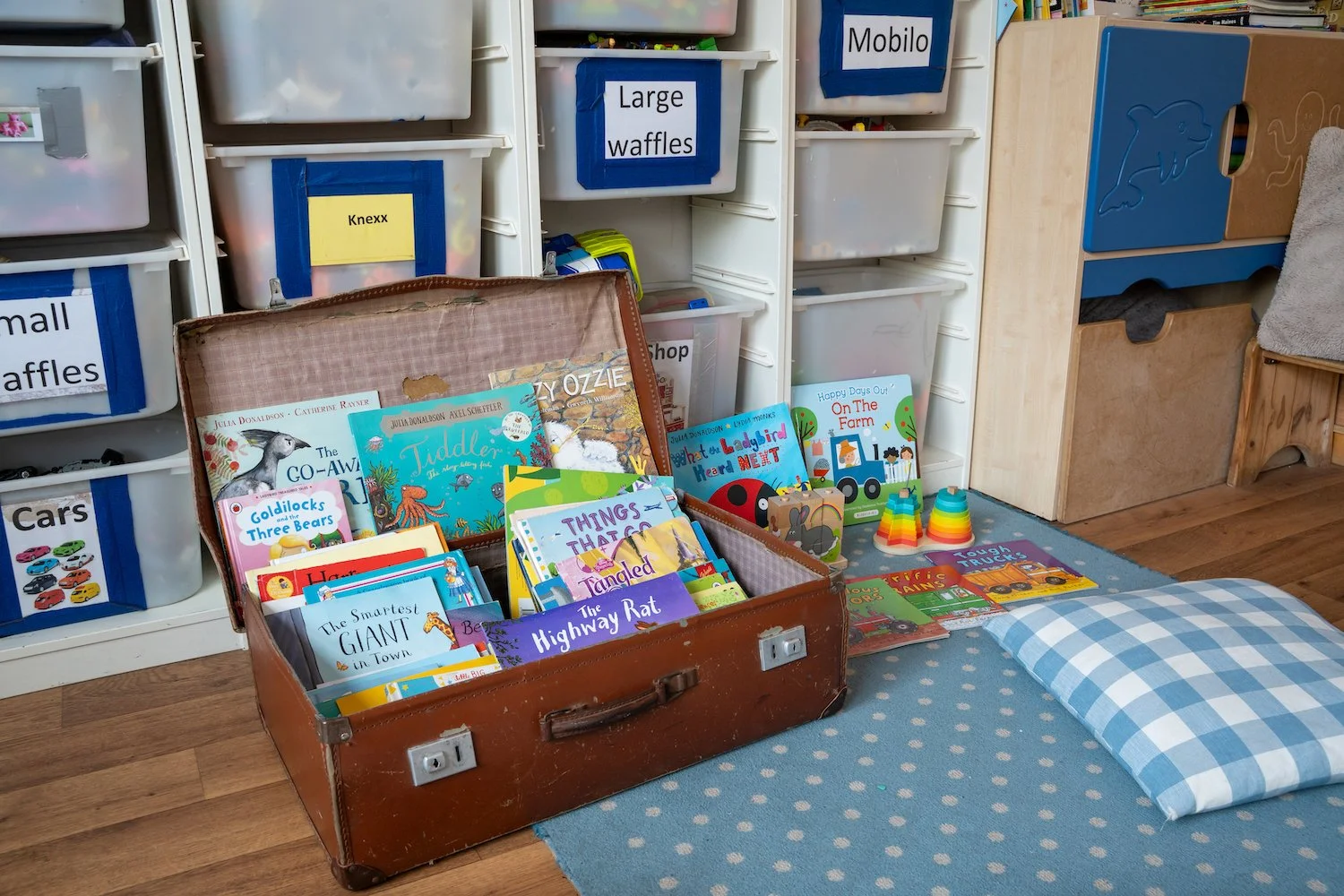 Children's library corner with a vintage suitcase filled with children's books, a blue and white checkered pillow, a blue rug, and plastic stacking cups on a child-sized table. Shelves in the background have labeled bins, including large waffles and 