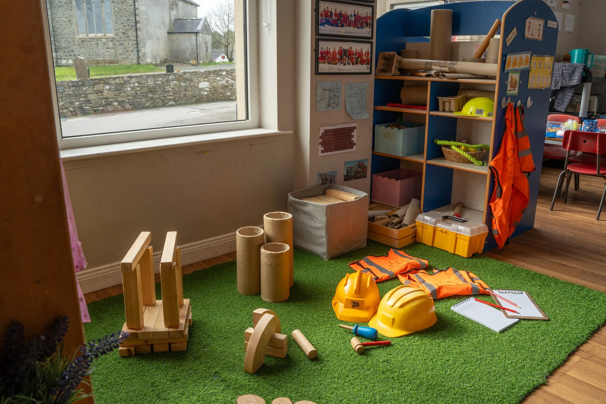 A play area inside a classroom with toy construction helmets, orange safety vests, wooden blocks, toy tools, a notebook, and shelves filled with educational materials, near a window showing an outdoor view.