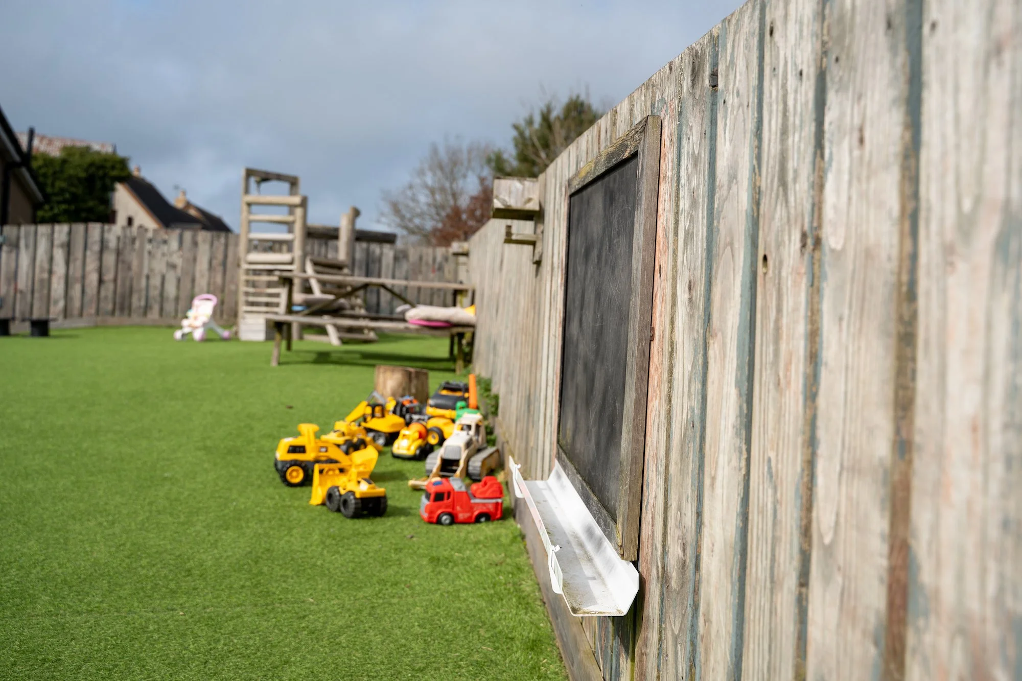An outdoor play area with a wooden fence, a chalkboard, and a row of children's toy cars on artificial grass, with outdoor furniture and a play structure in the background.