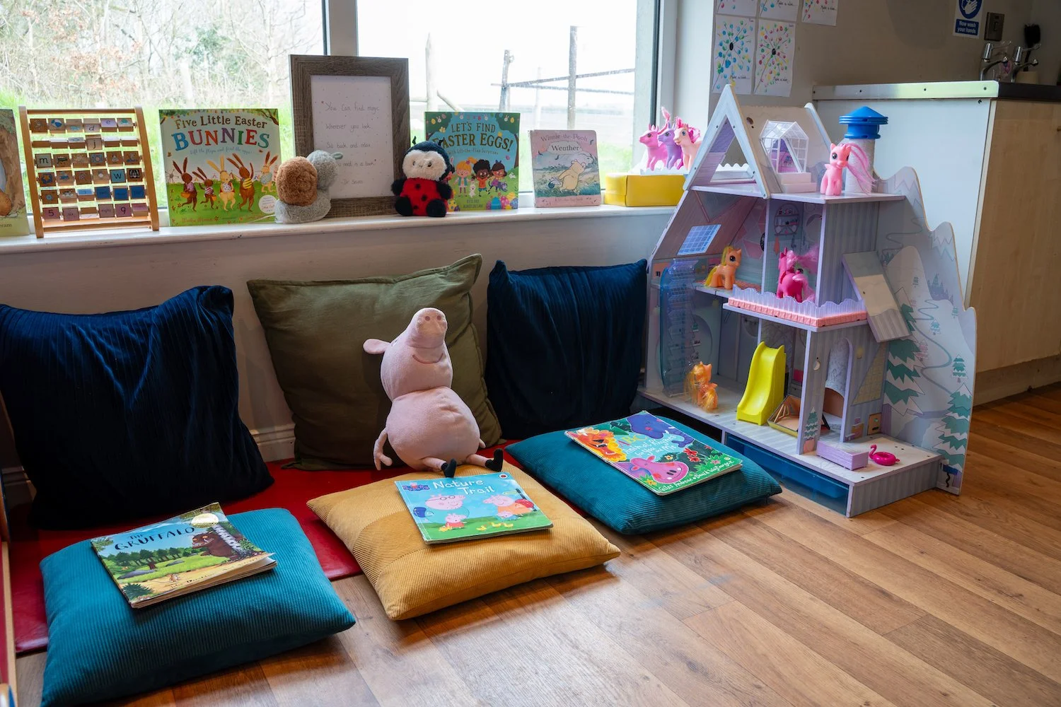 Cozy children's reading nook with colorful pillows, children's books, stuffed animals, and a dollhouse near a window with children's artwork and toys.
