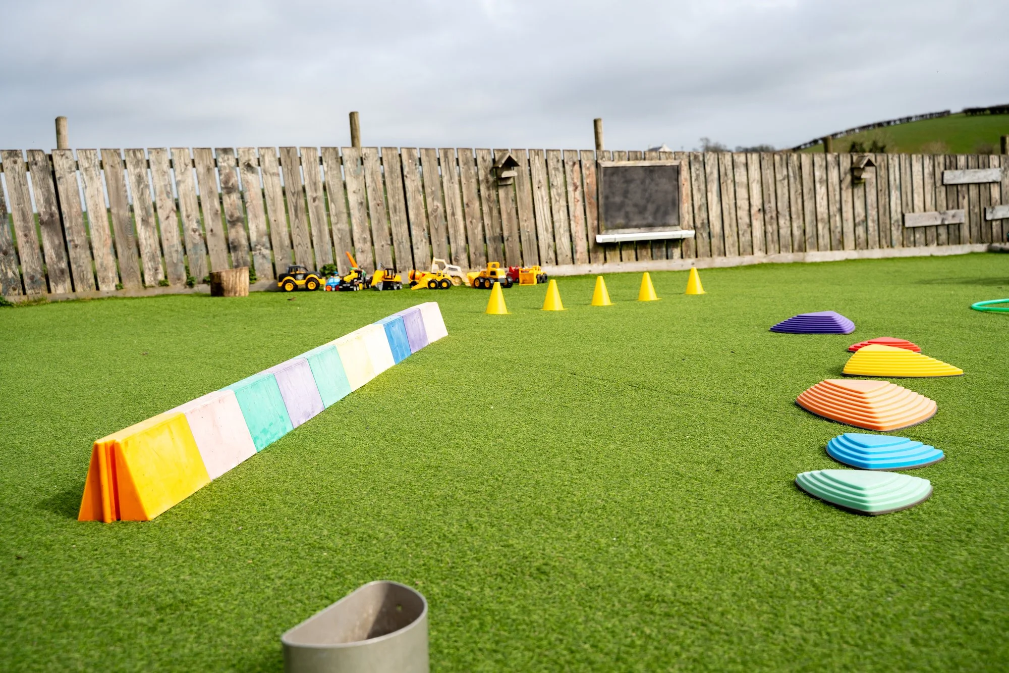 Outdoor play area with colorful plastic obstacle cones, toy trucks, and steps on artificial grass, enclosed by a wooden fence.