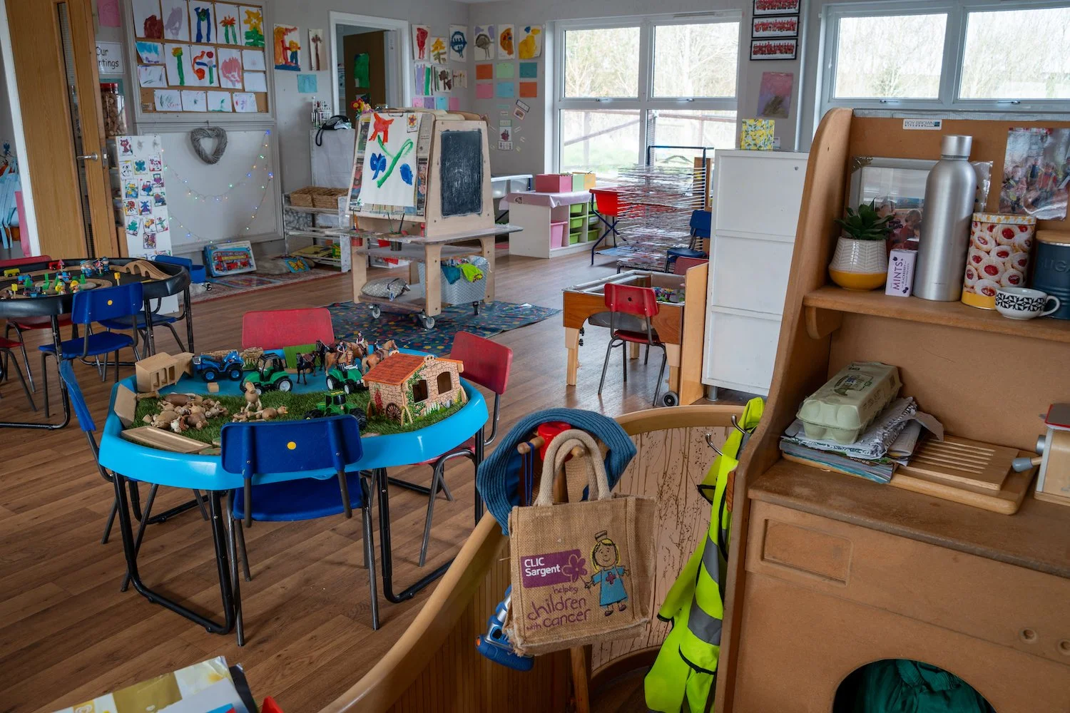 An indoor preschool classroom with colorful children's artwork on the walls, small tables with chairs, and various toys and educational materials visible throughout the room.
