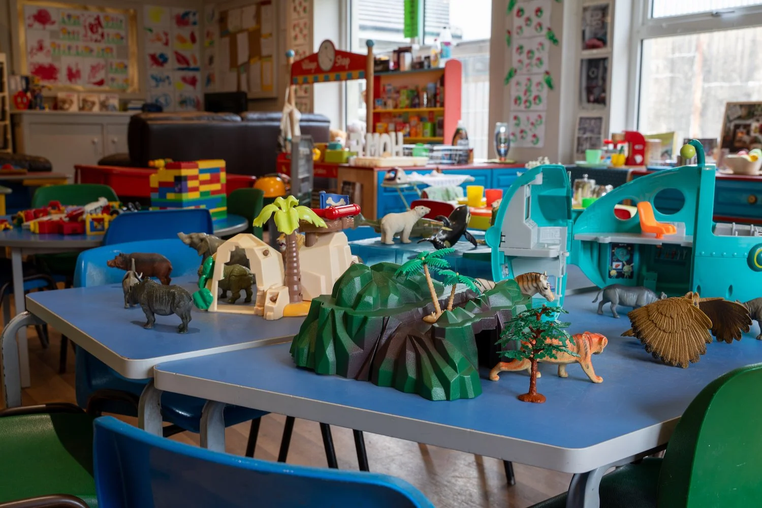 Children's playroom with blue tables and green chairs, featuring plastic animal figurines, jungle and mountain playsets, and a toy truck, with a colorful classroom background.