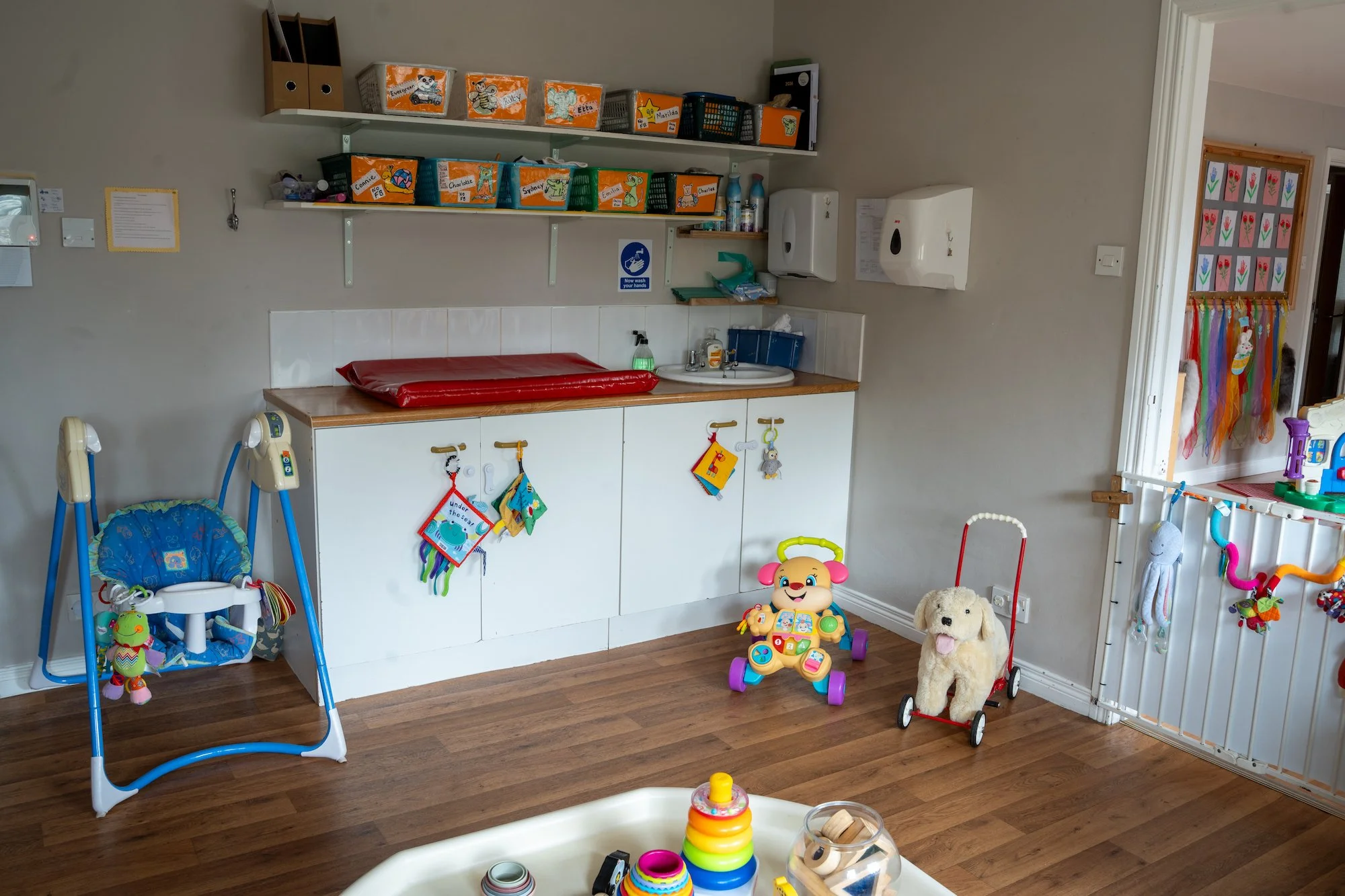 Kids' playroom with a changing station, toys, and a safety gate. Storage shelves on the wall hold labeled bins. Toys include a stuffed dog on a red wagon, a baby walker, and stacking rings on a table.