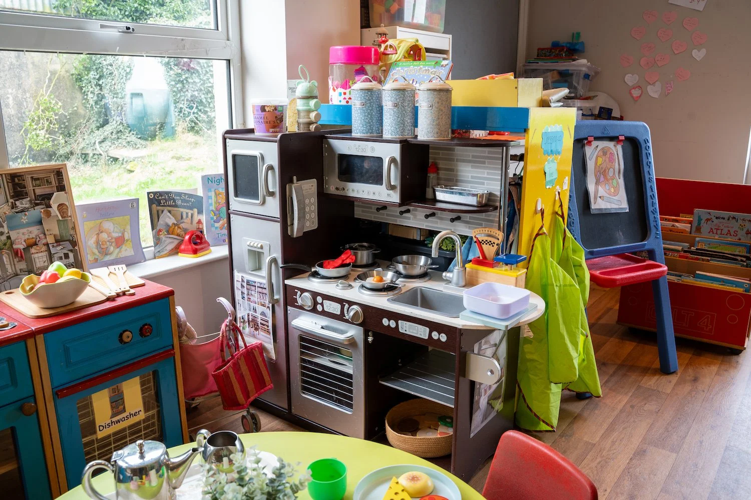 A cluttered play kitchen with toy stove, microwave, kitchen accessories, and children's books on a shelf, near a window.