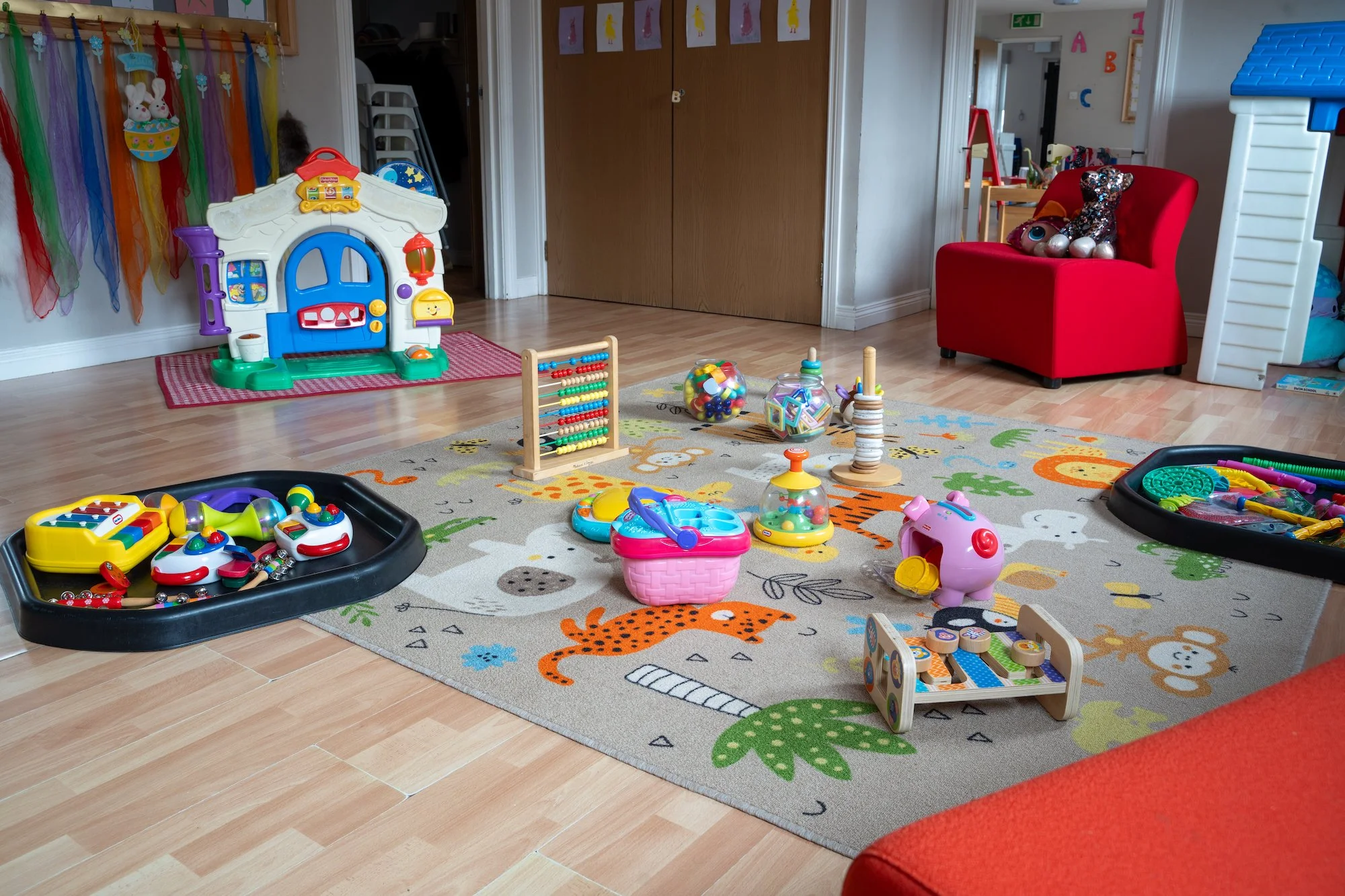 Colorful children's playroom with toys on a animal-themed rug, plastic playhouse, rocking horse, and various toys and decorations.