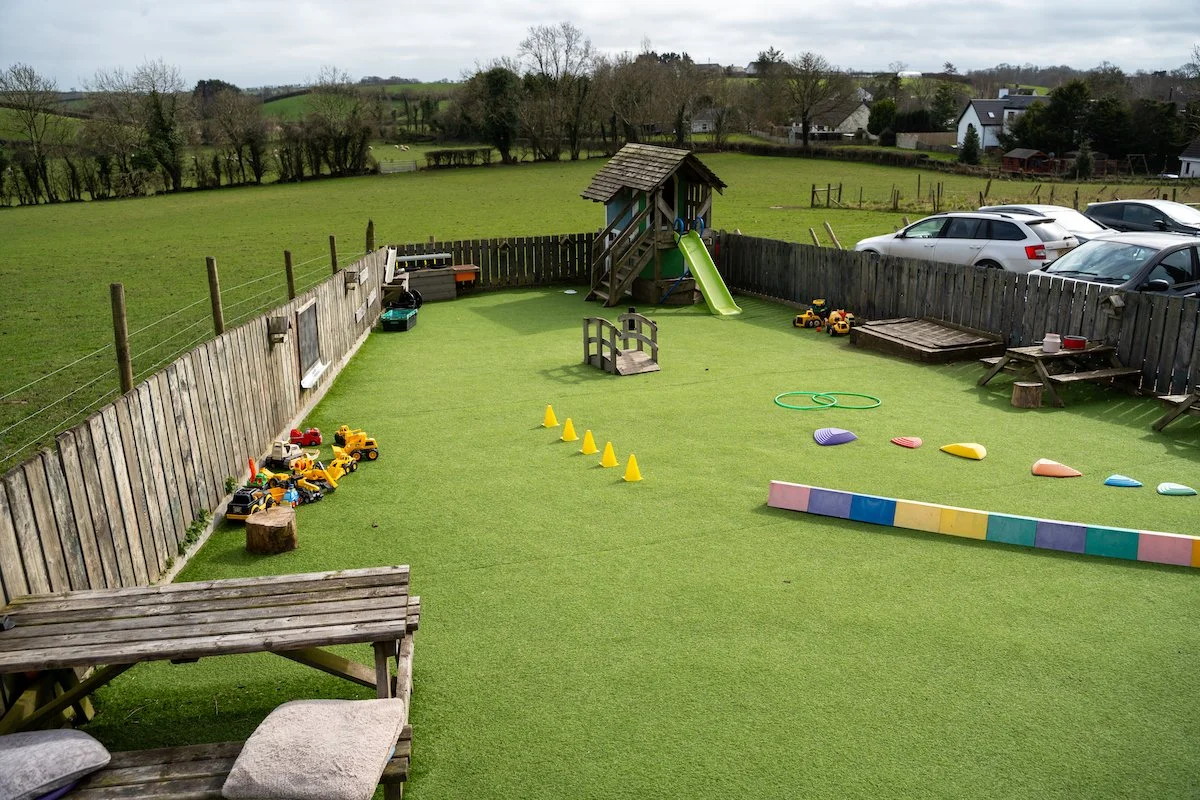 Empty children's outdoor play area with a small slide, toys, cones, and climbing ramps, surrounded by a wooden fence and neighboring houses with parking lot visible in the background.