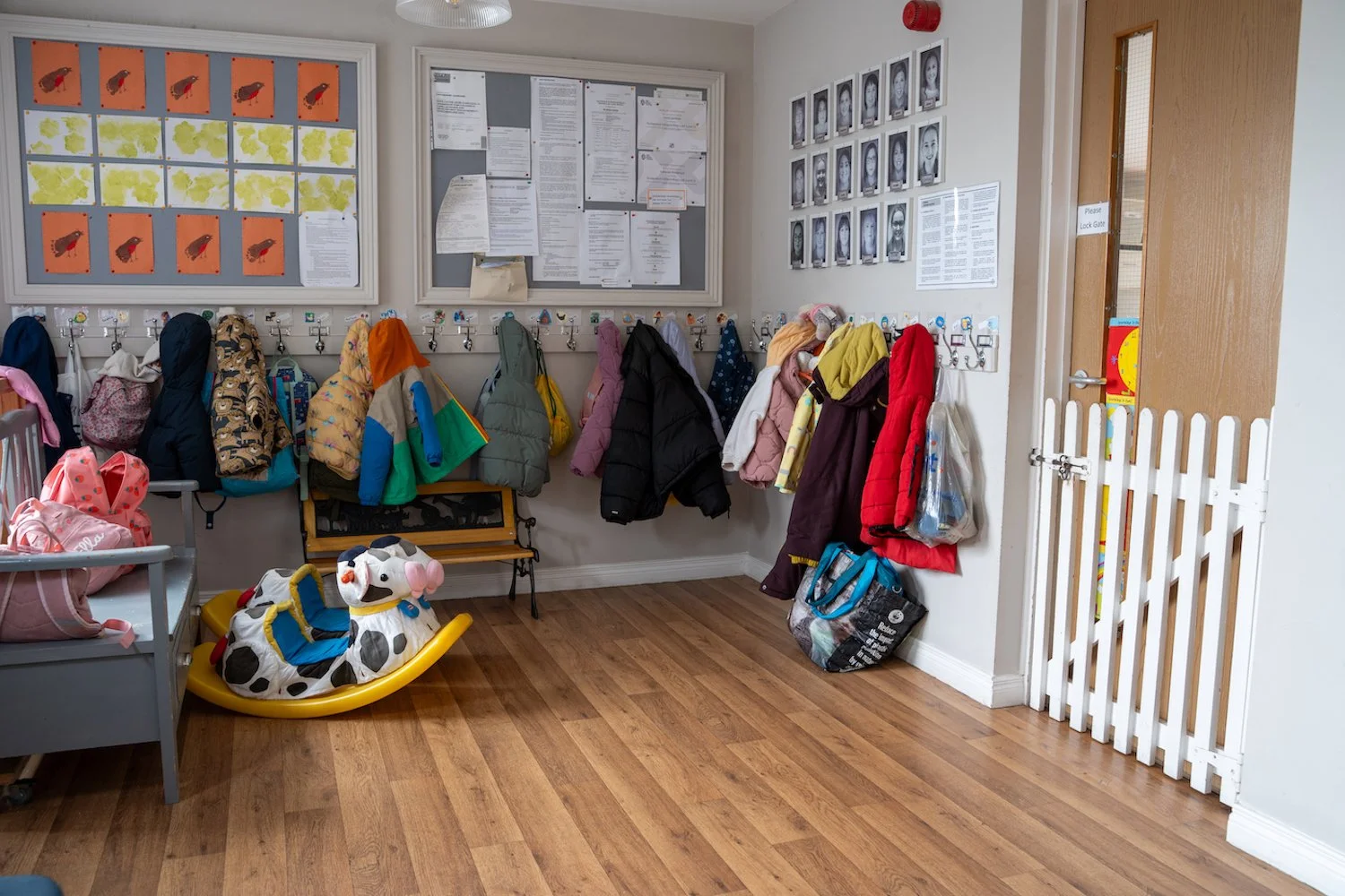 Coat hooks with jackets hanging, a dozen pairs of shoes, a frog shape rocker, bulletin boards with notices and photos in a daycare or preschool entryway.