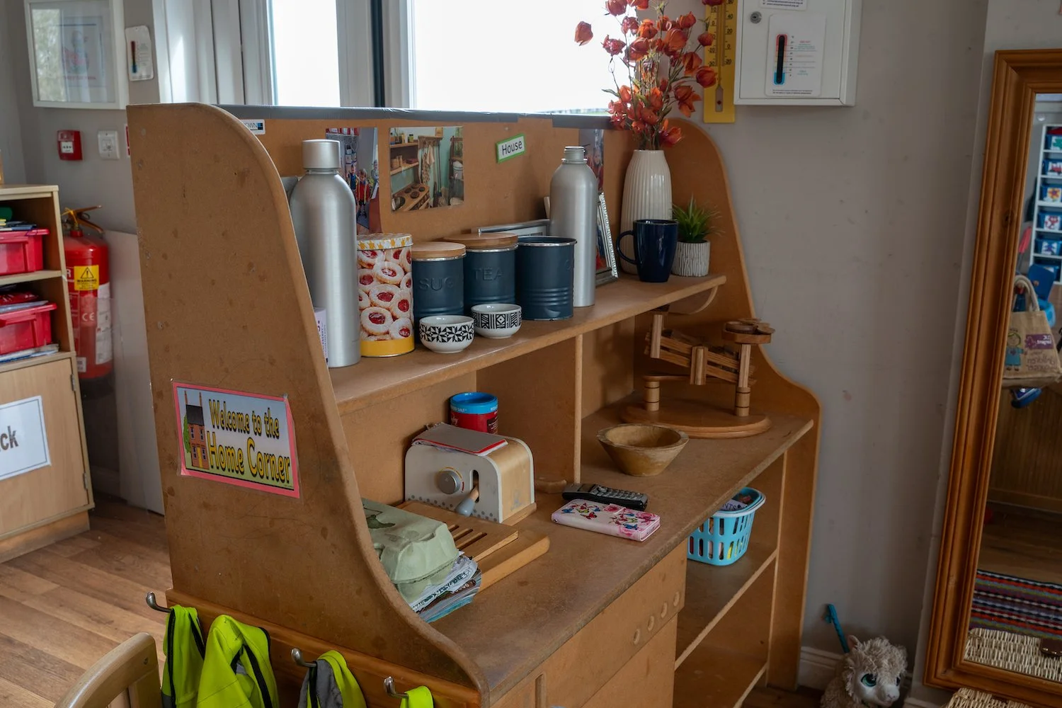 A wooden organization shelf with a sign saying "Welcome to the Home Corner." The shelf holds cans of tea and sugar, a couple of bottles, a small potted plant, bowls, and a wooden platform. Below are jackets hanging, and nearby are a remote, a cloth, 
