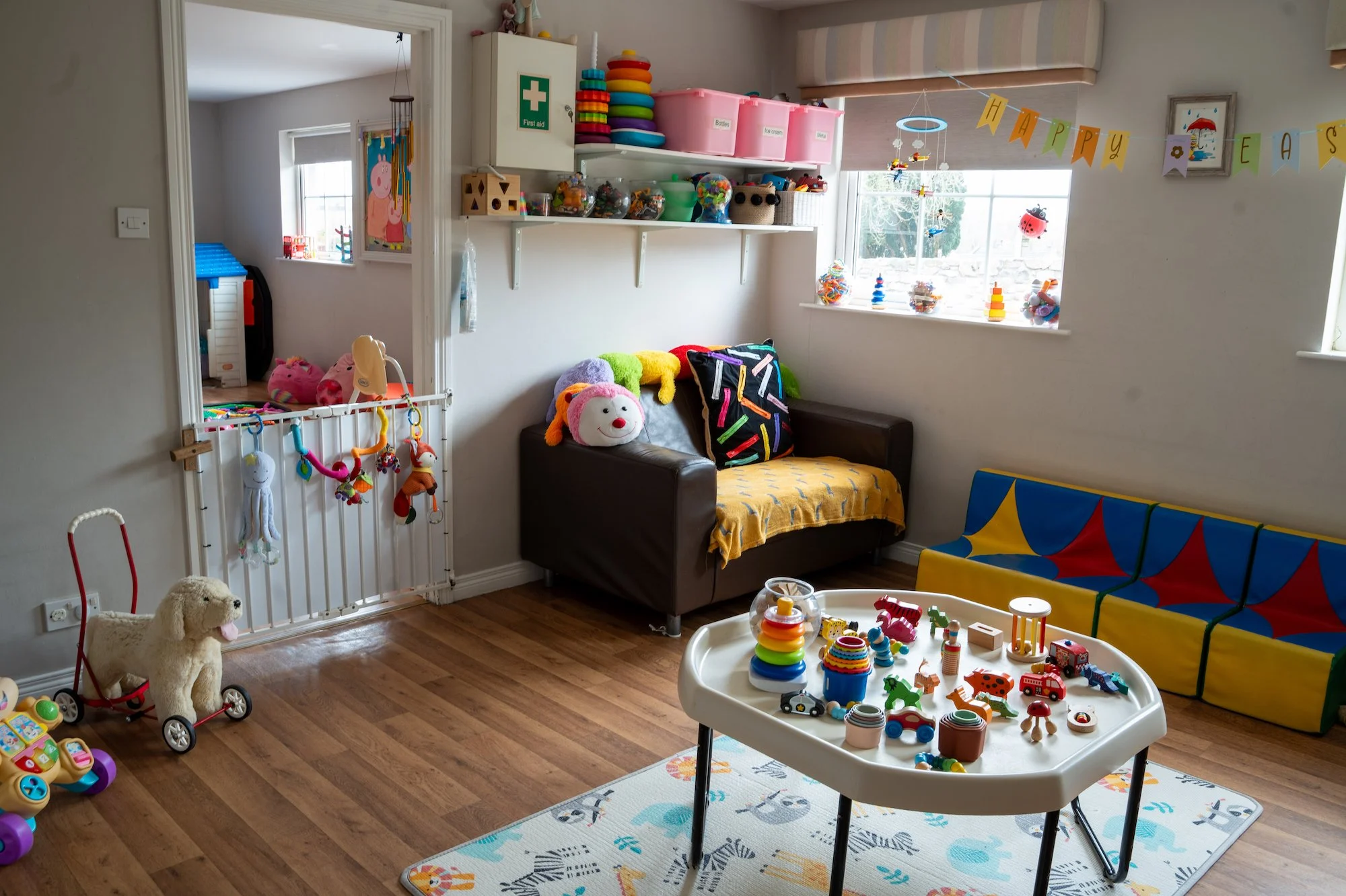 Children's playroom with toys on a table, stuffed animals on a sofa, a playpen with hanging toys, colorful cushions, and a banner reading 'Happy Easter'.