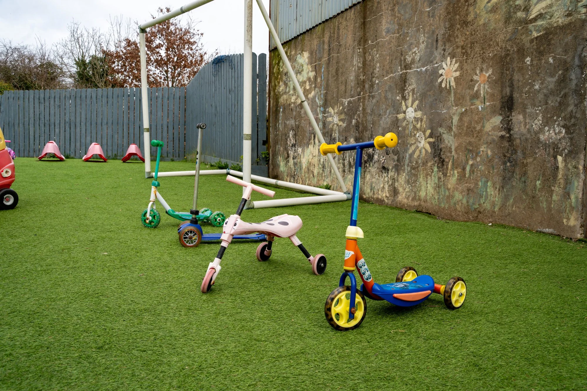 Children's outdoor play area with small scooters and a toy car on a grassy field, enclosed by a blue wooden fence and a weathered wall with painted flowers.