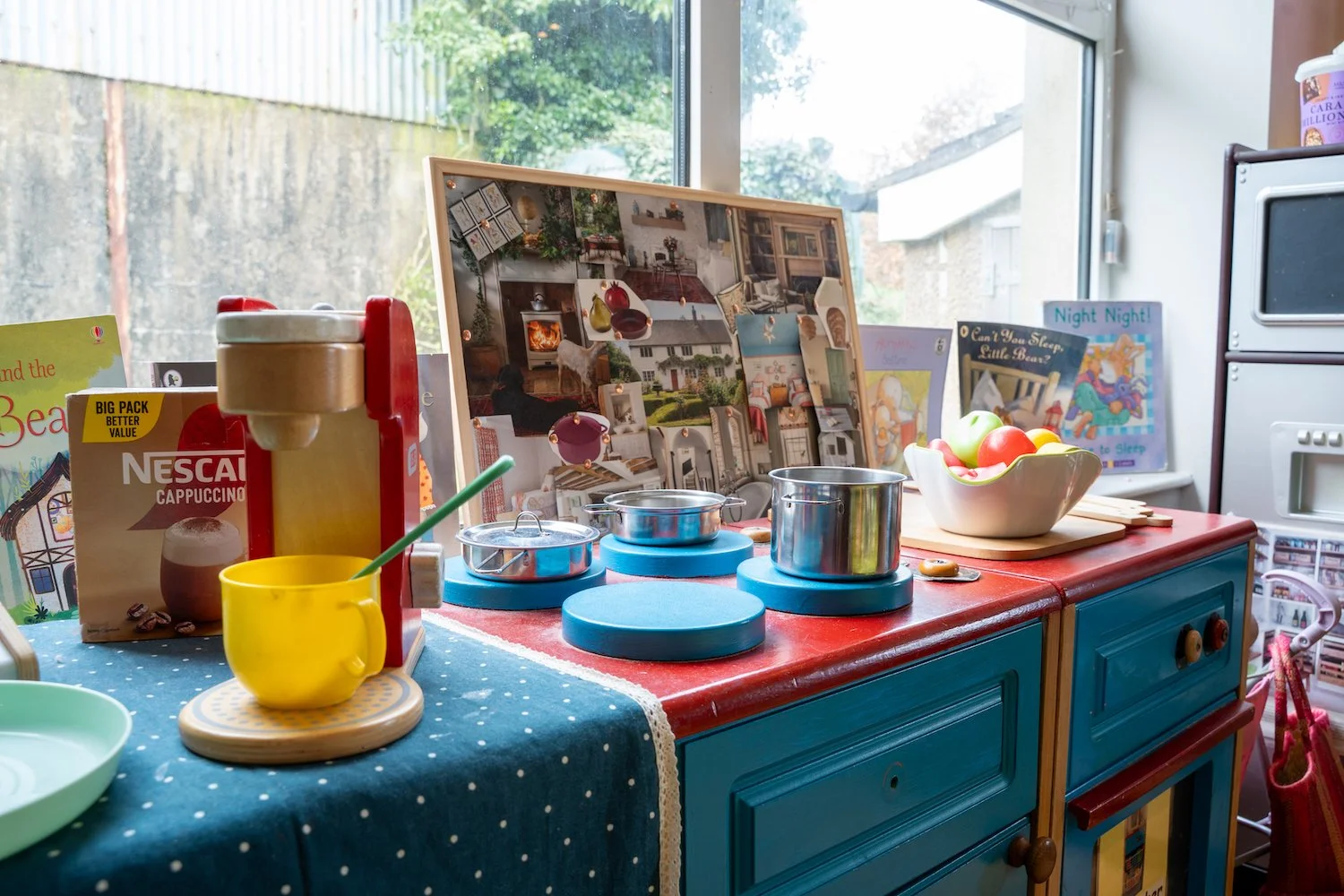 Children's play kitchen with toy pots, bowls, and food items, set against a window with a backyard view.