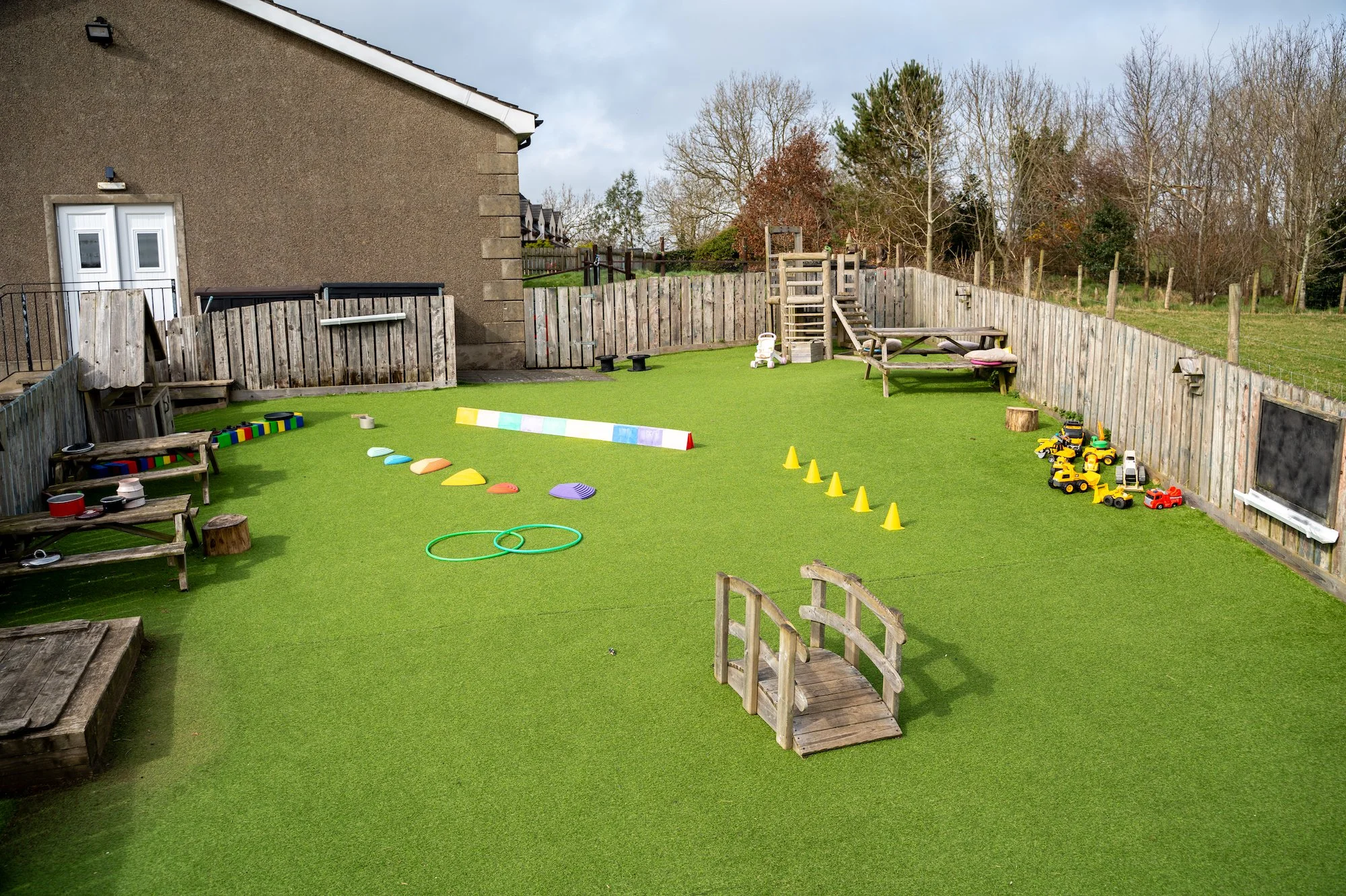 Children's outdoor play area with artificial grass, wooden fences, and various toys including toy trucks, cones, hoops, and climbing structures, set against a building and trees in the background.