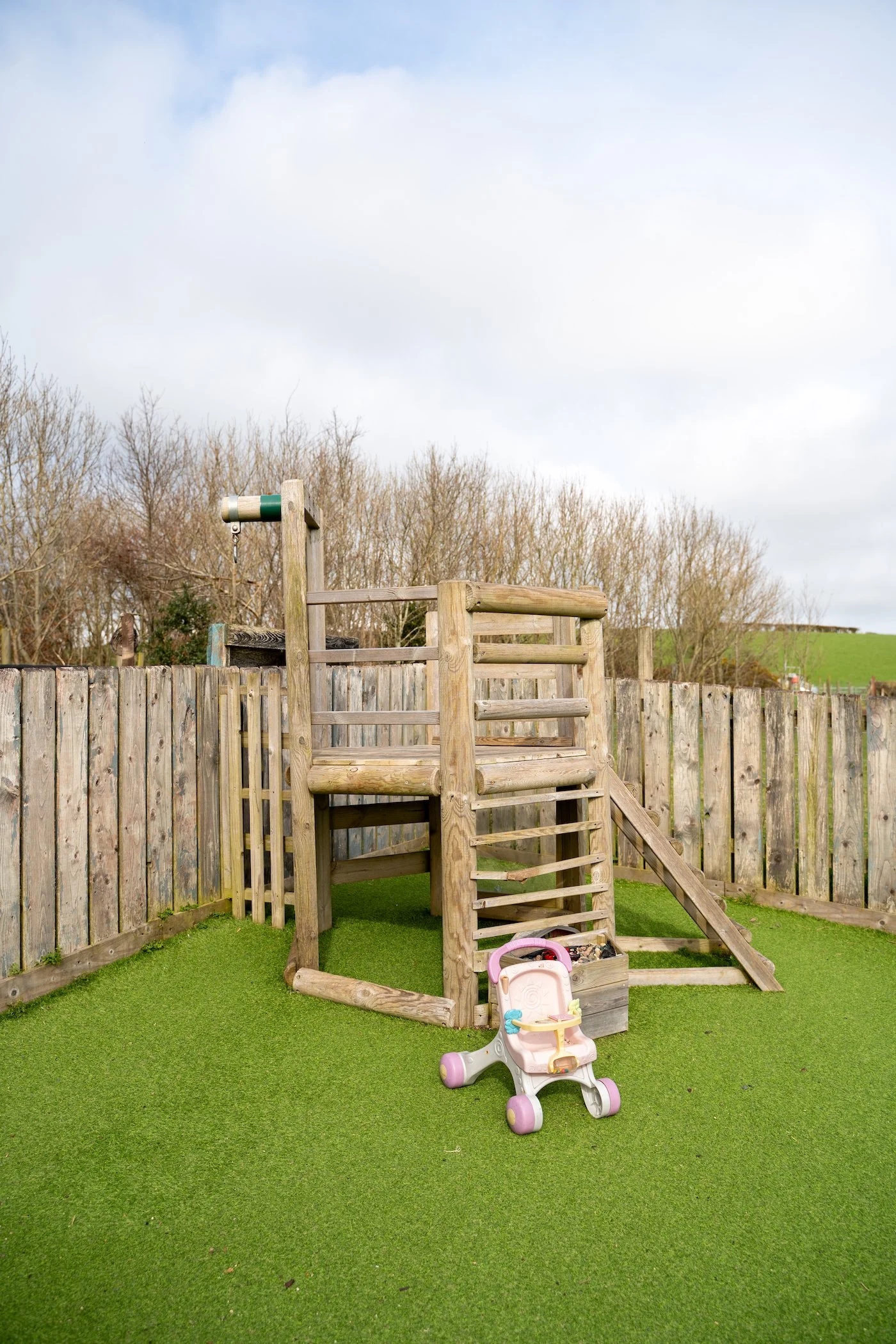 A wooden play structure with ladder and slide, a pink and white toy stroller, and a fence in a backyard with green grass.