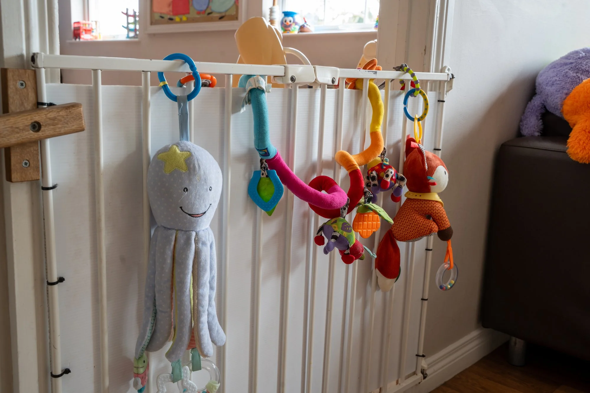 A white baby crib with hanging plush toys, including an octopus, a teething ring, a ladybug, and a fox, next to a purple pillow on a black couch in a room with a window.