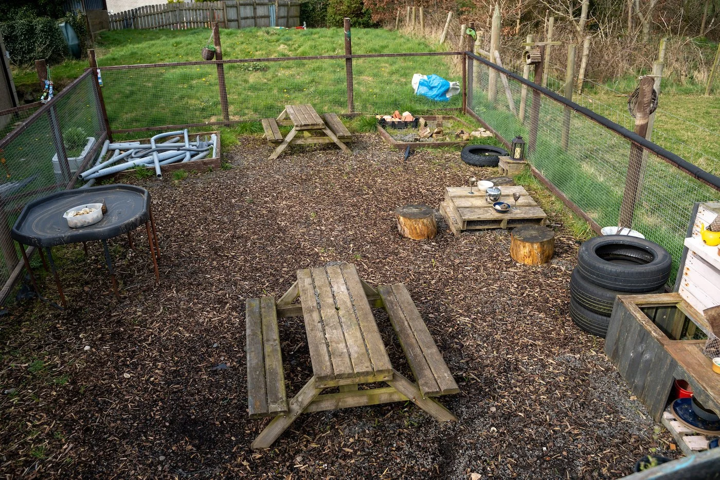Backyard with leaf mulch ground, picnic table, wooden pallets, tires, and a fenced area with grass.