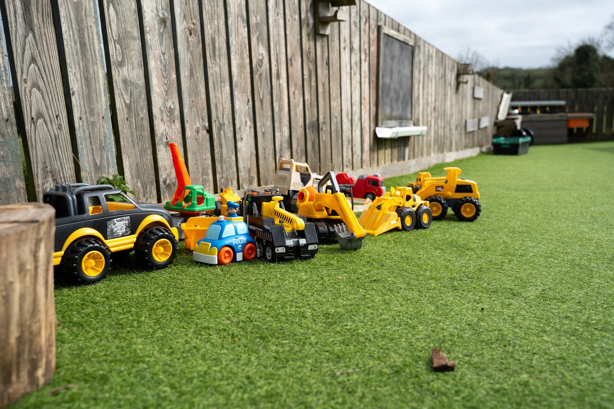 Line of children's toy construction vehicles and cars on grass beside a wooden fence.