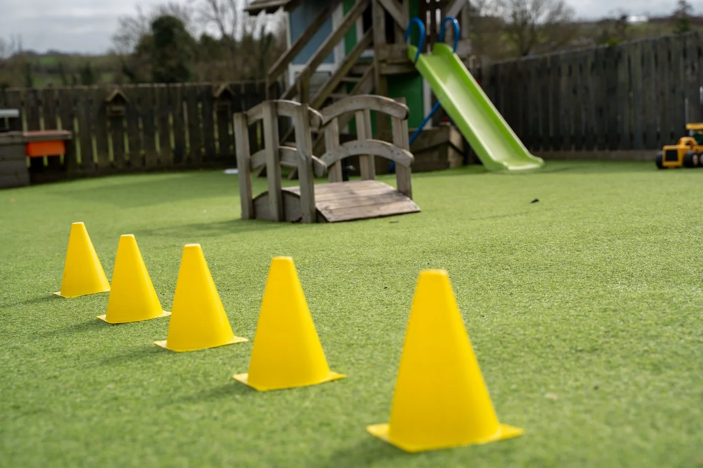 Playground with green artificial turf, yellow traffic cones arranged in a line, a small wooden bridge, and a slide in the background.