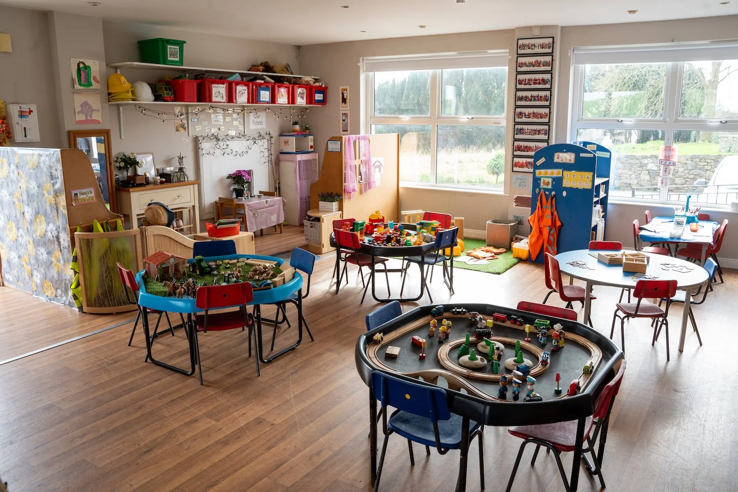 Bright kindergarten classroom with tables of toys, building blocks, a train set, and art supplies; windows showing an outdoor view.