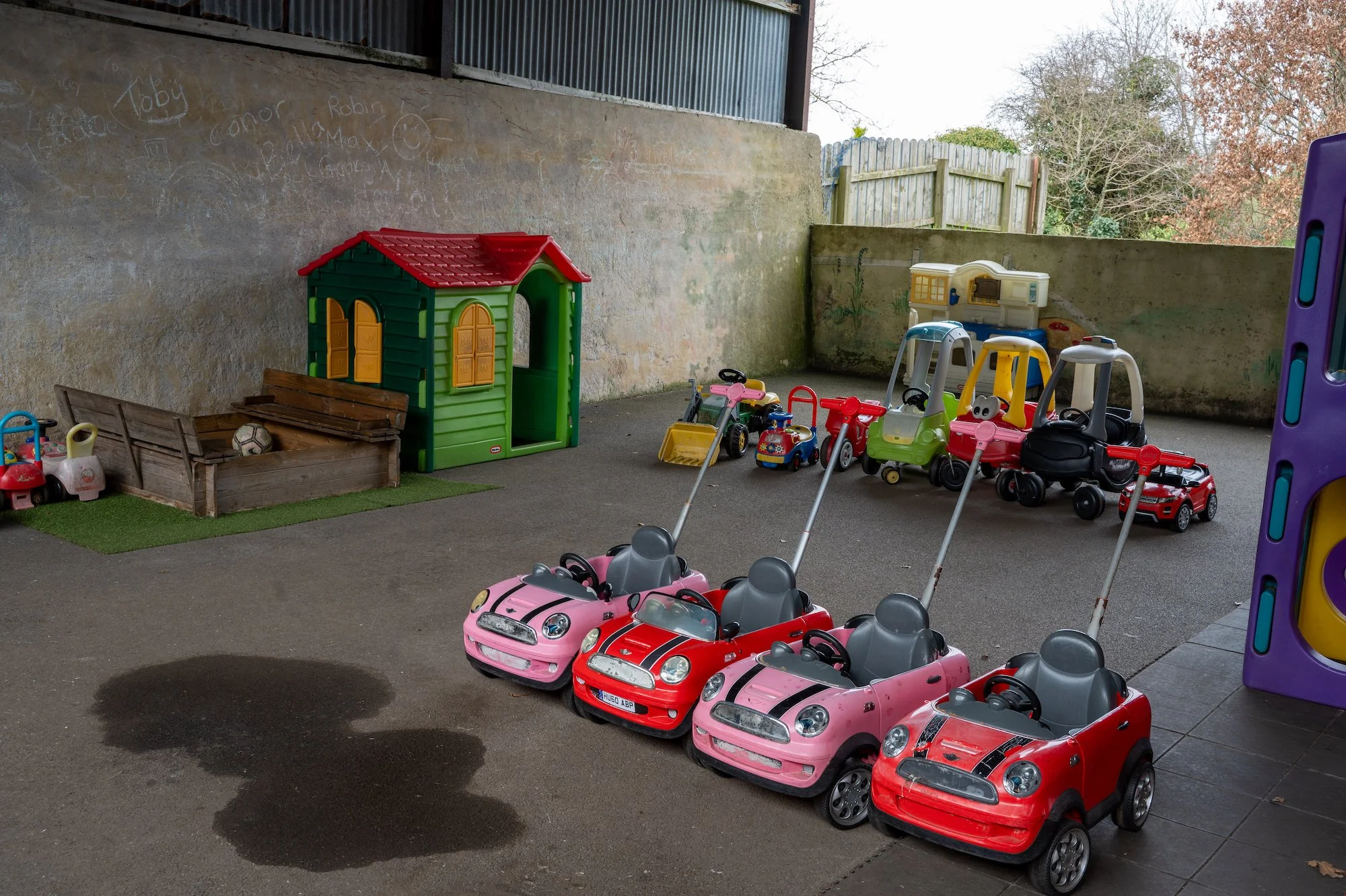 Play area with small toy cars, a green playhouse, and various toys including a sandbox with sand toys, a plastic slide, and a small playhouse on a patio.