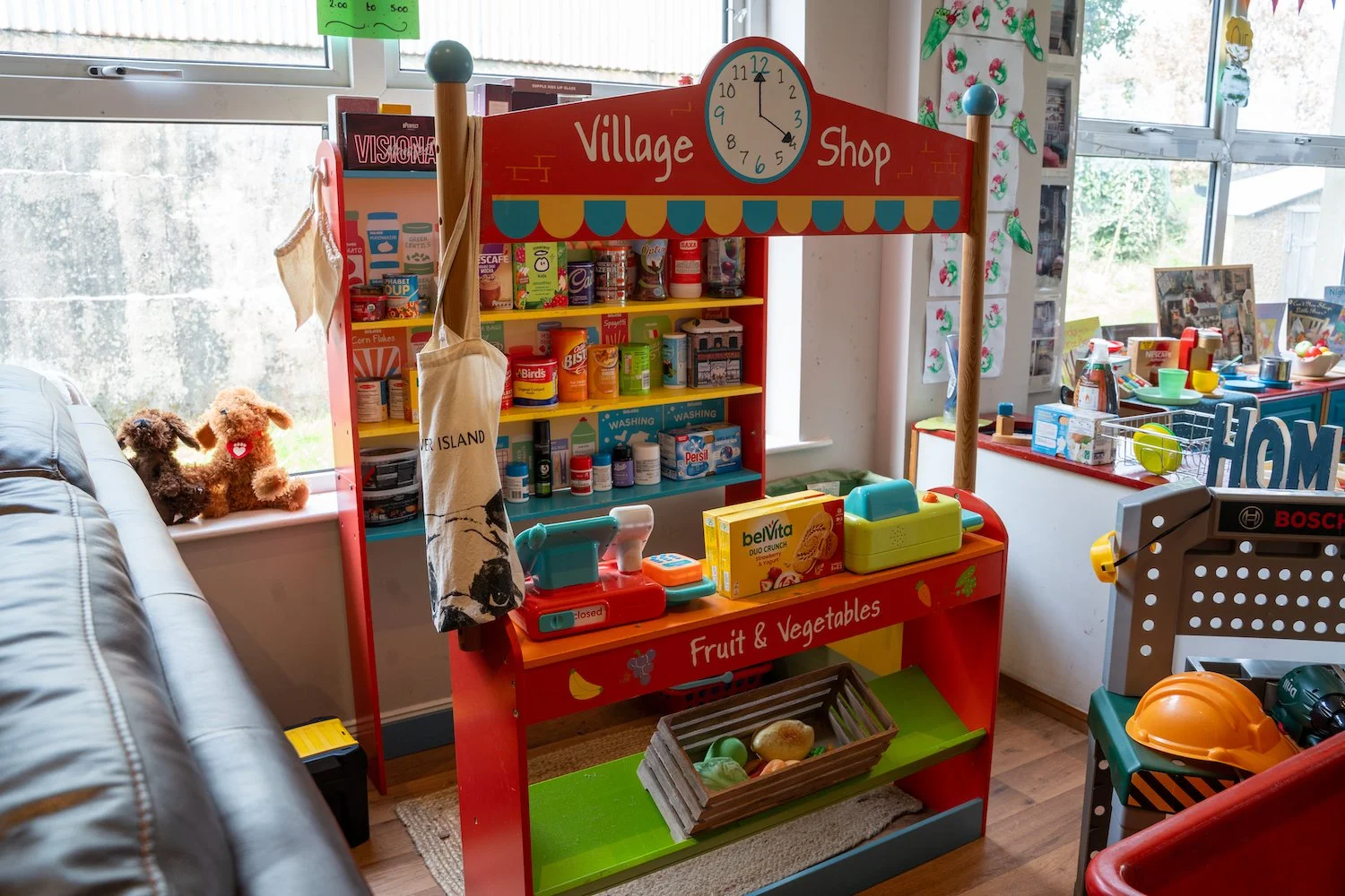Indoor pretend play Village Shop area with shelves of toy food and supplies, a shopping cart, and plush toys on the window sill.