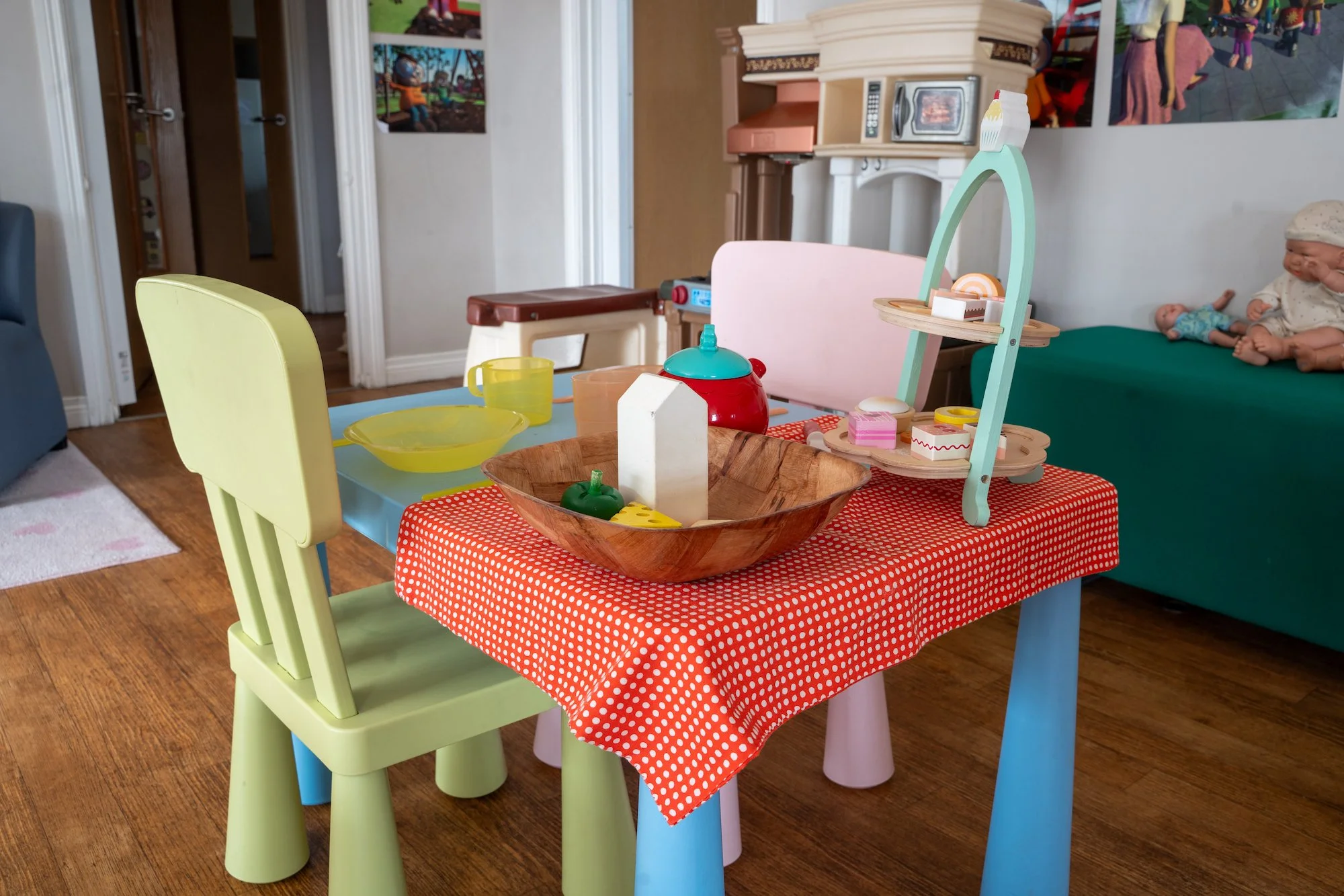 Children's playroom with small table covered in red polka dot tablecloth, set with wooden bowl, toy food, and play kitchen utensils, surrounded by colorful chairs, with a dark green couch and wall posters in the background.
