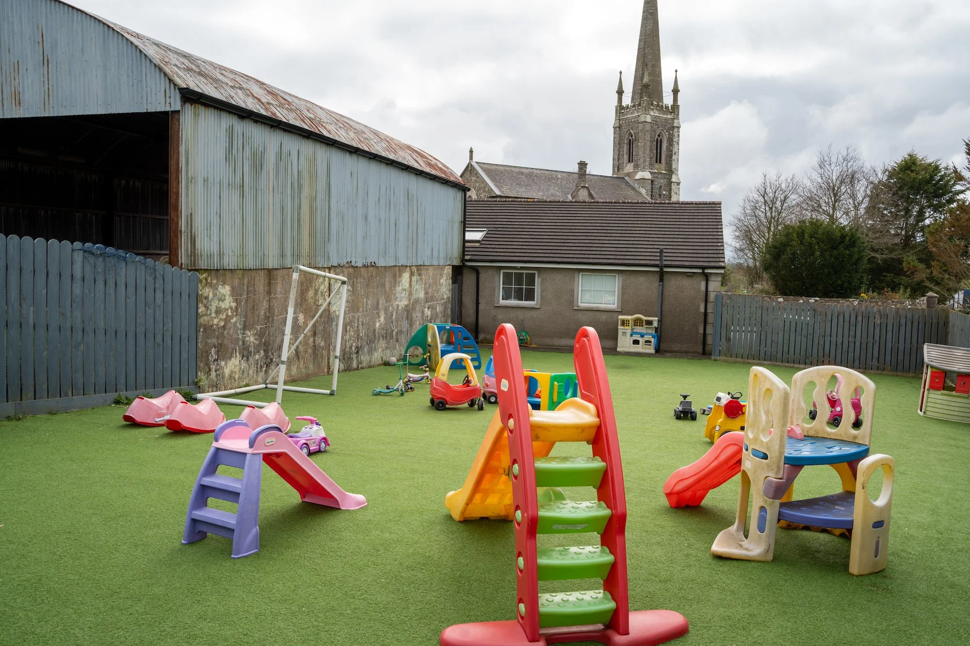 Children's playground with small slides, a toy car, and other toys on artificial grass, surrounded by fences and buildings, with a church in the background.