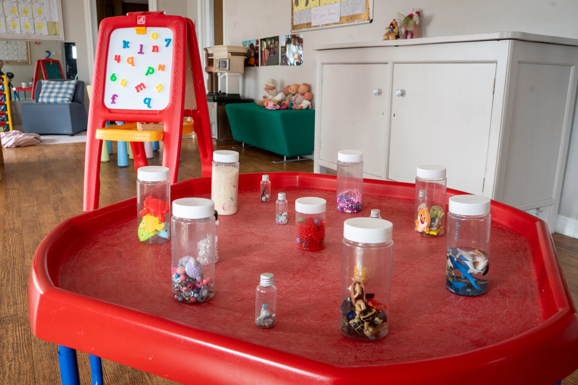 A red plastic table with various jars filled with beads, sequins, and small toys. In the background, a whiteboard with colorful alphabet magnets and a red and white easel with magnetic letters.