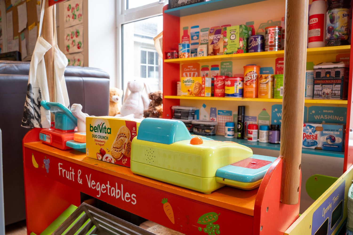 A colorful children's play food stall labeled 'Fruit & Vegetables' with toy kitchen appliances and toy food items, including a toy oven and a toy cash register, set inside a store or play area.