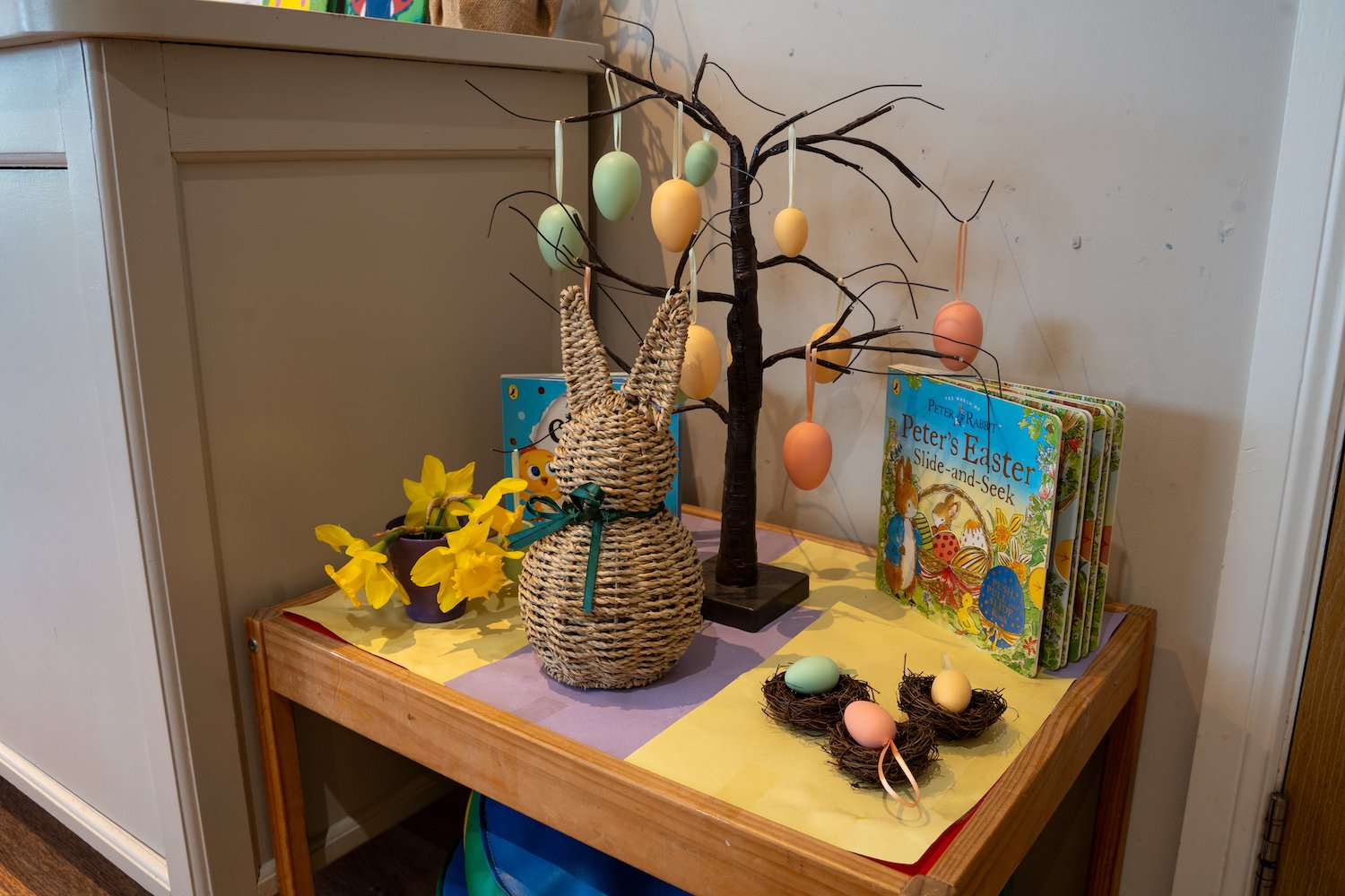 Easter-themed decorations on a wooden table including a small decorated Easter tree with hanging eggs, a woven rabbit figure, a pot of yellow daffodils, colorful Easter books, and small nests with eggs.