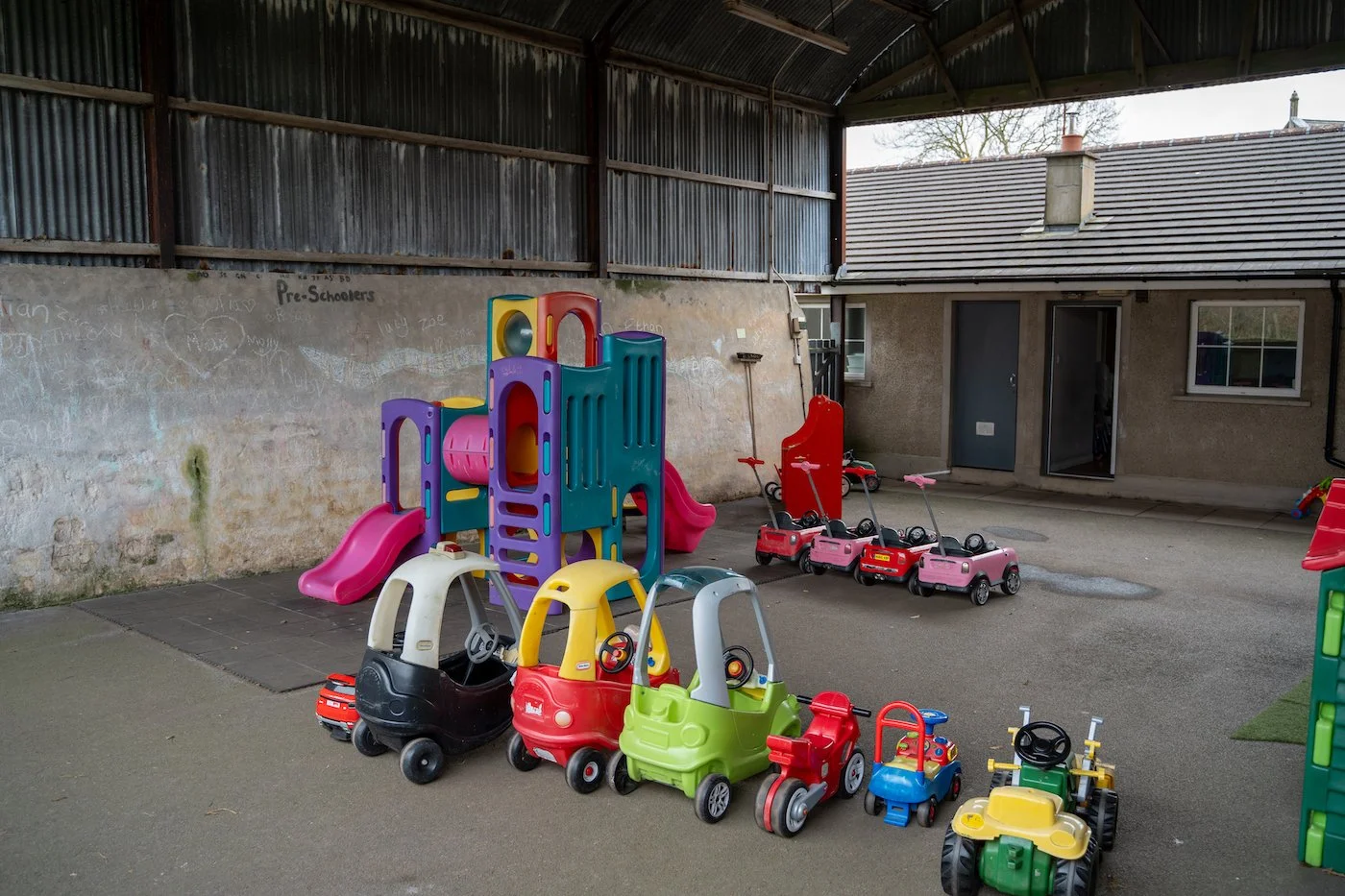 An outdoor covered play area with various colorful plastic and toy vehicles and slides, set against a wall with chalk drawings and a building in the background.