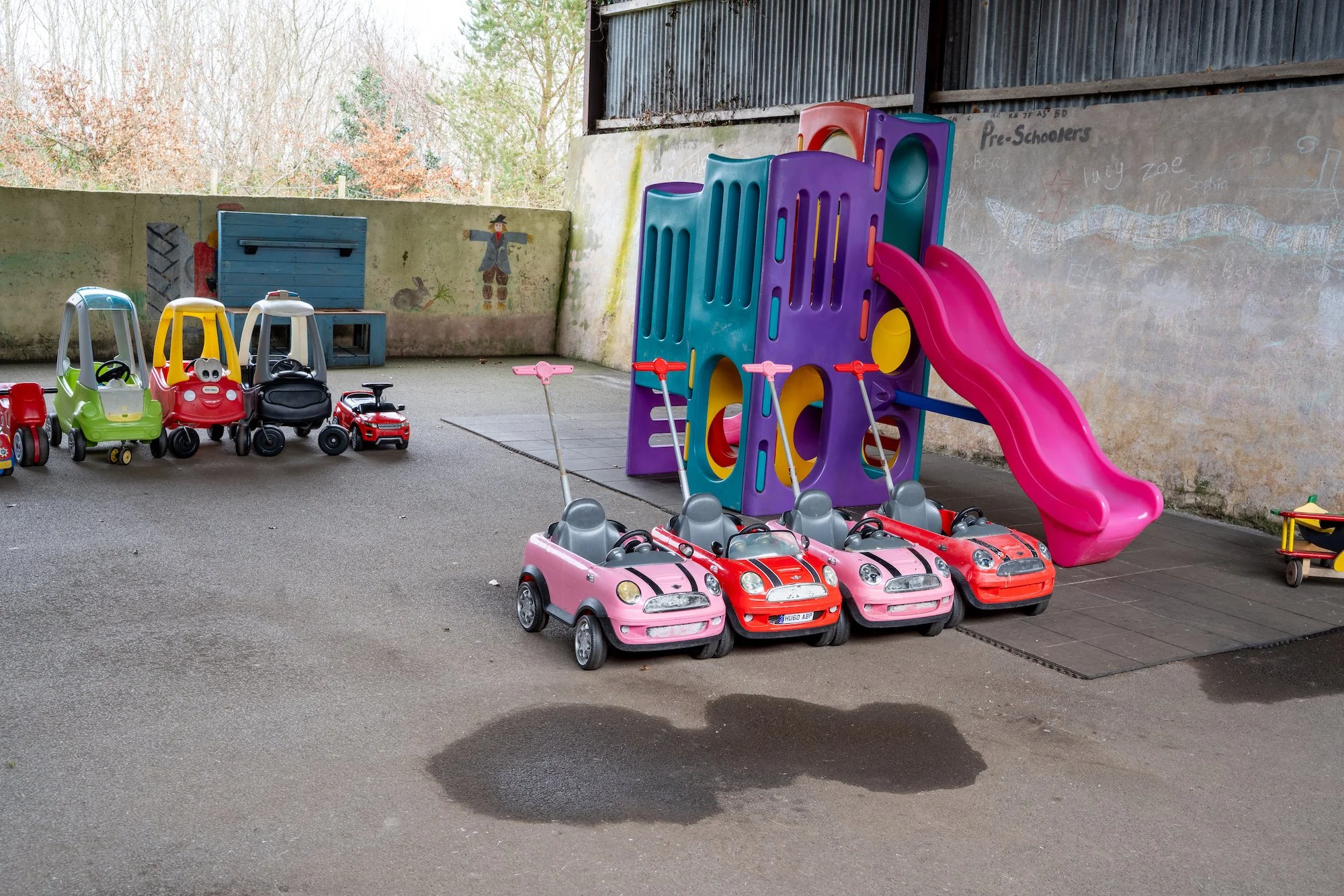 Children's playground with colorful toy cars, a purple and pink slide, and a concrete wall with child drawings in an outdoor setting.