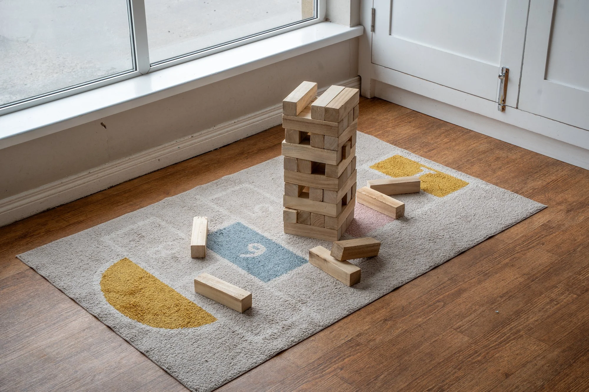 A Jenga game stacked with wooden blocks on a rug with colorful shapes, near a glass door in a room with hardwood flooring.