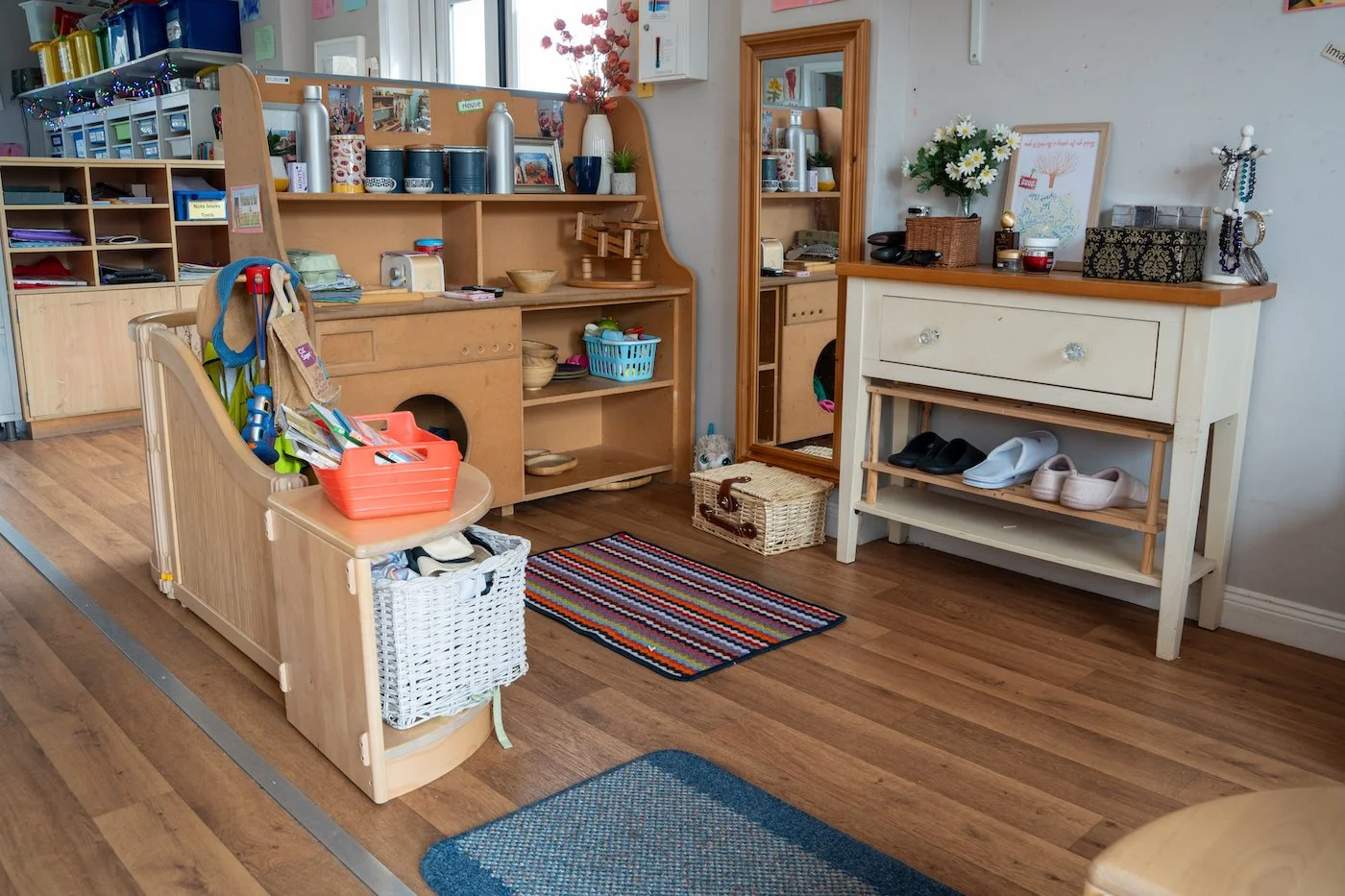 Interior of a classroom or daycare with wooden furniture, shelves, baskets, and various supplies, including shoes and toys.