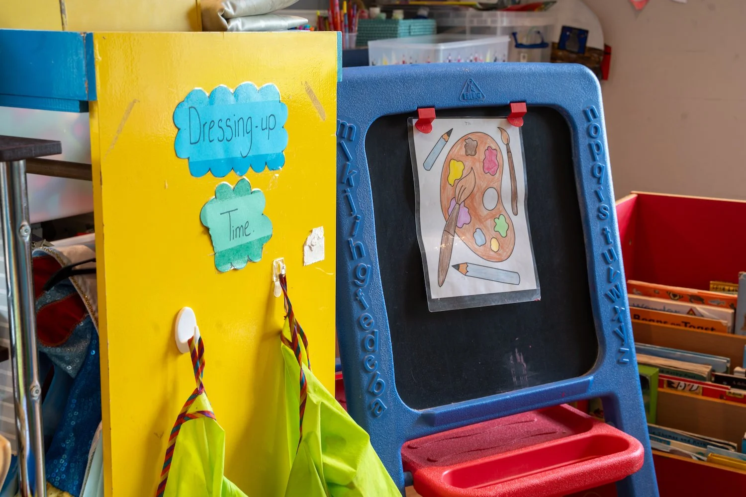 A yellow display board with blue cloud-shaped signs labeled 'Dressing up' and 'Time,' with two yellow and one green apron hanging from white hooks. Next to it is a blue plastic easel with a paper of a colorful artist's palette and brushes attached wi