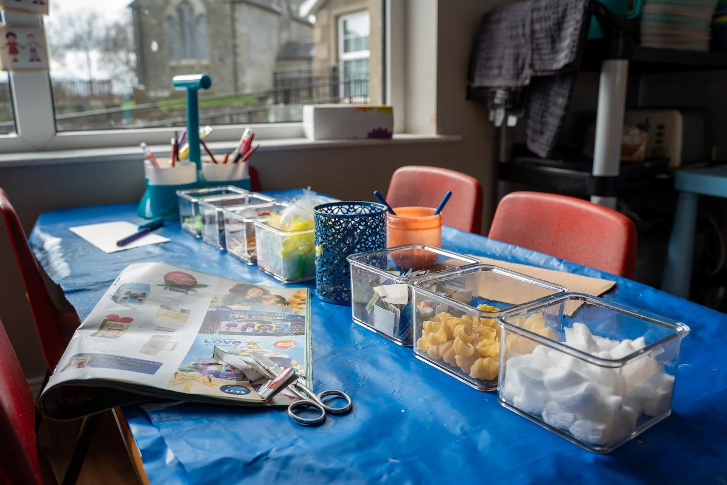 A table covered with a blue plastic tablecloth holds various craft supplies in clear containers, scissors, and a newspaper with scissors on top. In the background, there is a window with a view of buildings outside, and several red chairs are around 