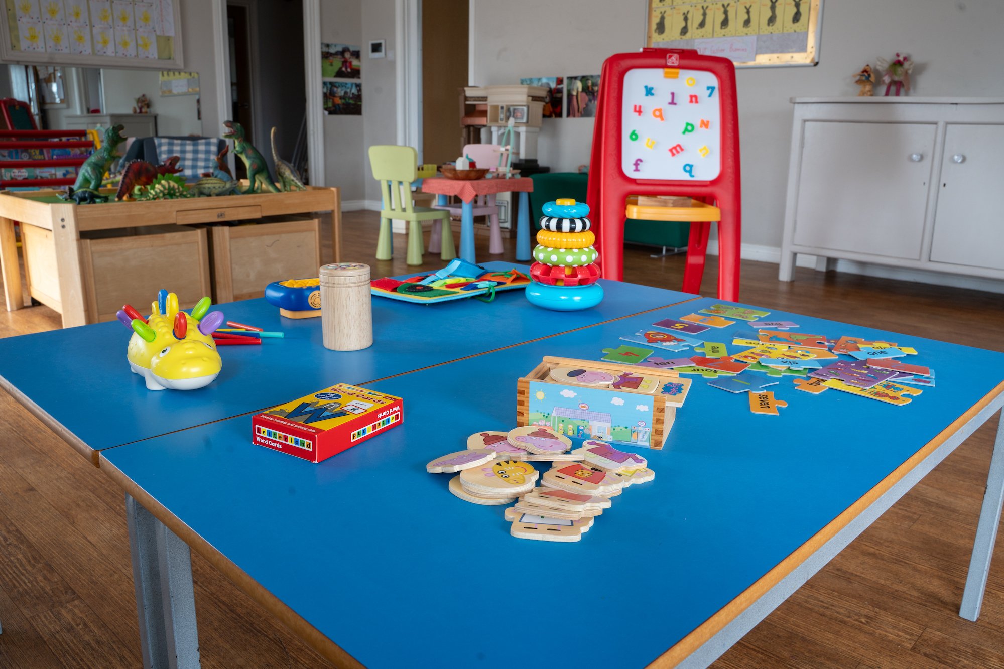 Children's playroom with a blue table filled with colorful toys including stacking rings, word and picture cards, and a yellow dinosaur-shaped plush; in the background, there are dinosaur figurines, a small table with chairs, and drawing boards.