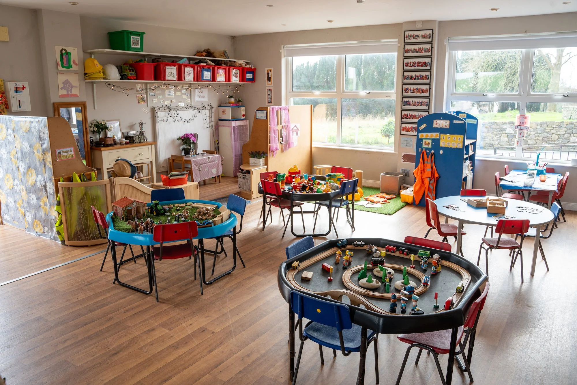 A brightly lit preschool classroom with various tables filled with toys, including a train set, blocks, and small figurines. There are window views outside, educational posters on the walls, and storage bins for classroom materials.