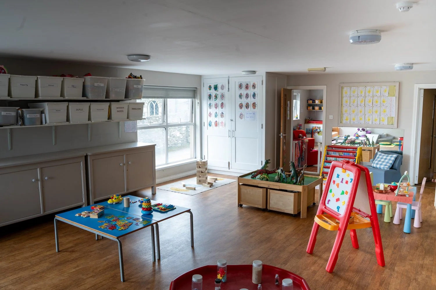A colorful preschool or daycare classroom with toys, play tables, storage bins, and artwork on the walls.