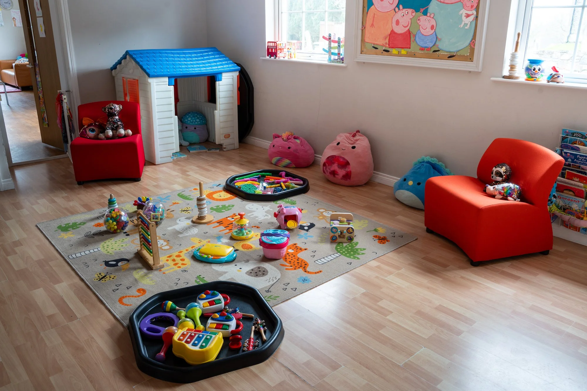 A children's playroom with toys scattered on a colorful animal-themed rug. There are two red chairs, one with plush toys including a Peppa Pig figure, and several plush bean bag toys against the wall. A small plastic playhouse and a tray of toys are also visible, with books on a bookshelf near the window. The room is brightly lit by natural light from the window.