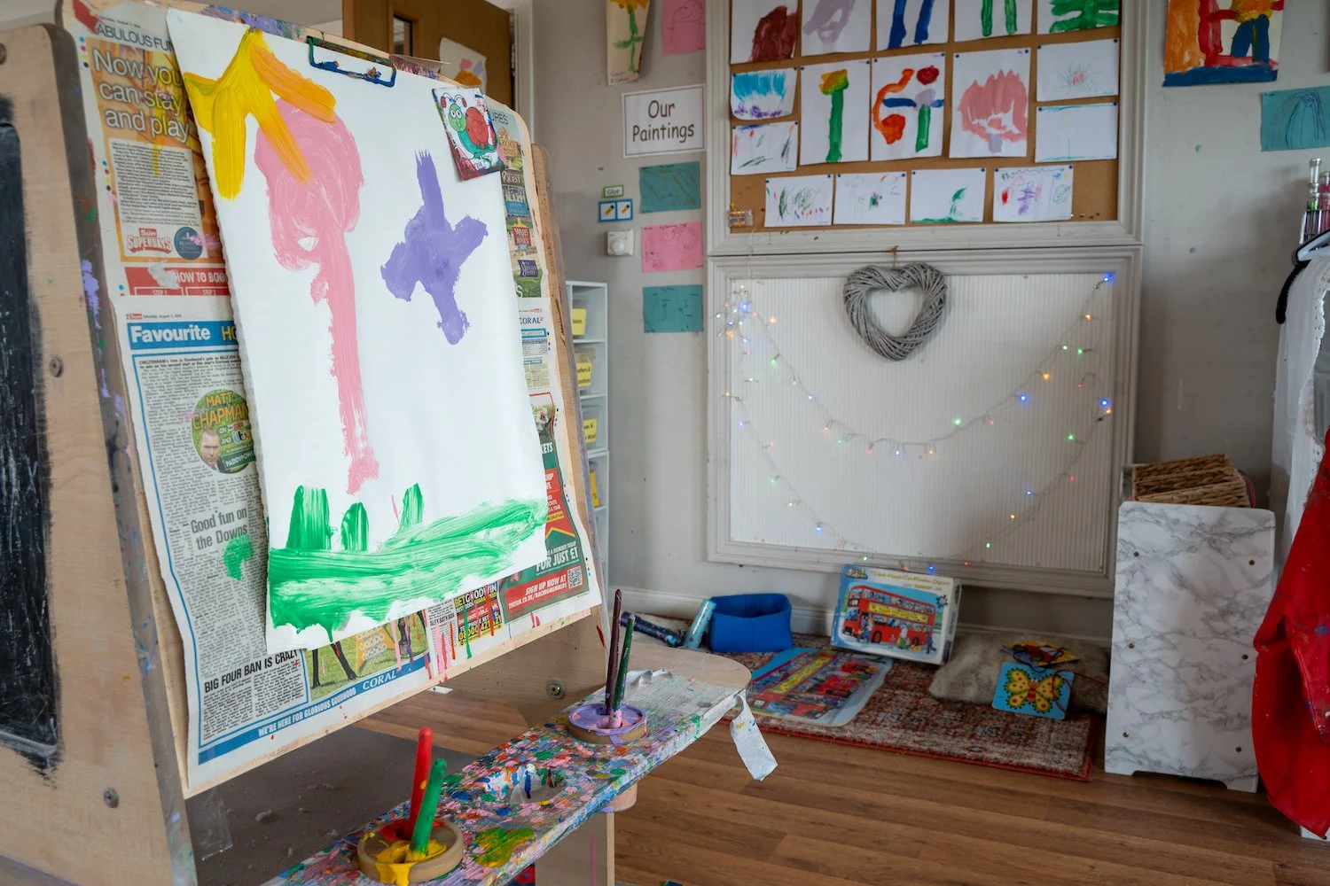 Children's art classroom with a large whiteboard displaying painted artwork, posters, and children's drawings on the wall, fairy lights, a heart-shaped wreath, and various art supplies and books on the floor and shelves.