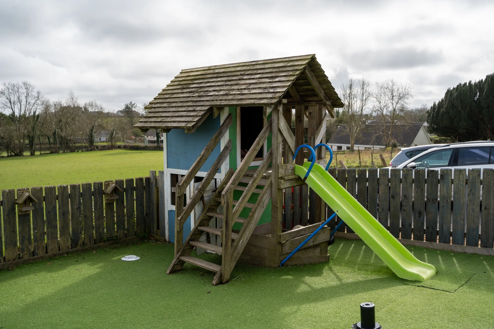 A small wooden playhouse with a green slide in a fenced backyard. The playhouse has a slanted roof and a staircase leading to its entrance. The yard has artificial grass, and there are some cars parked outside the fence, with trees and houses in the 